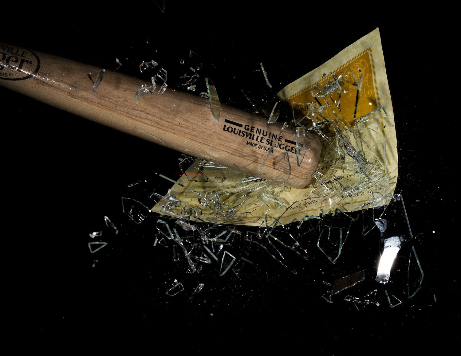 A wooden Louisville Slugger baseball bat smashes through paper money with flying glass shards on black background.