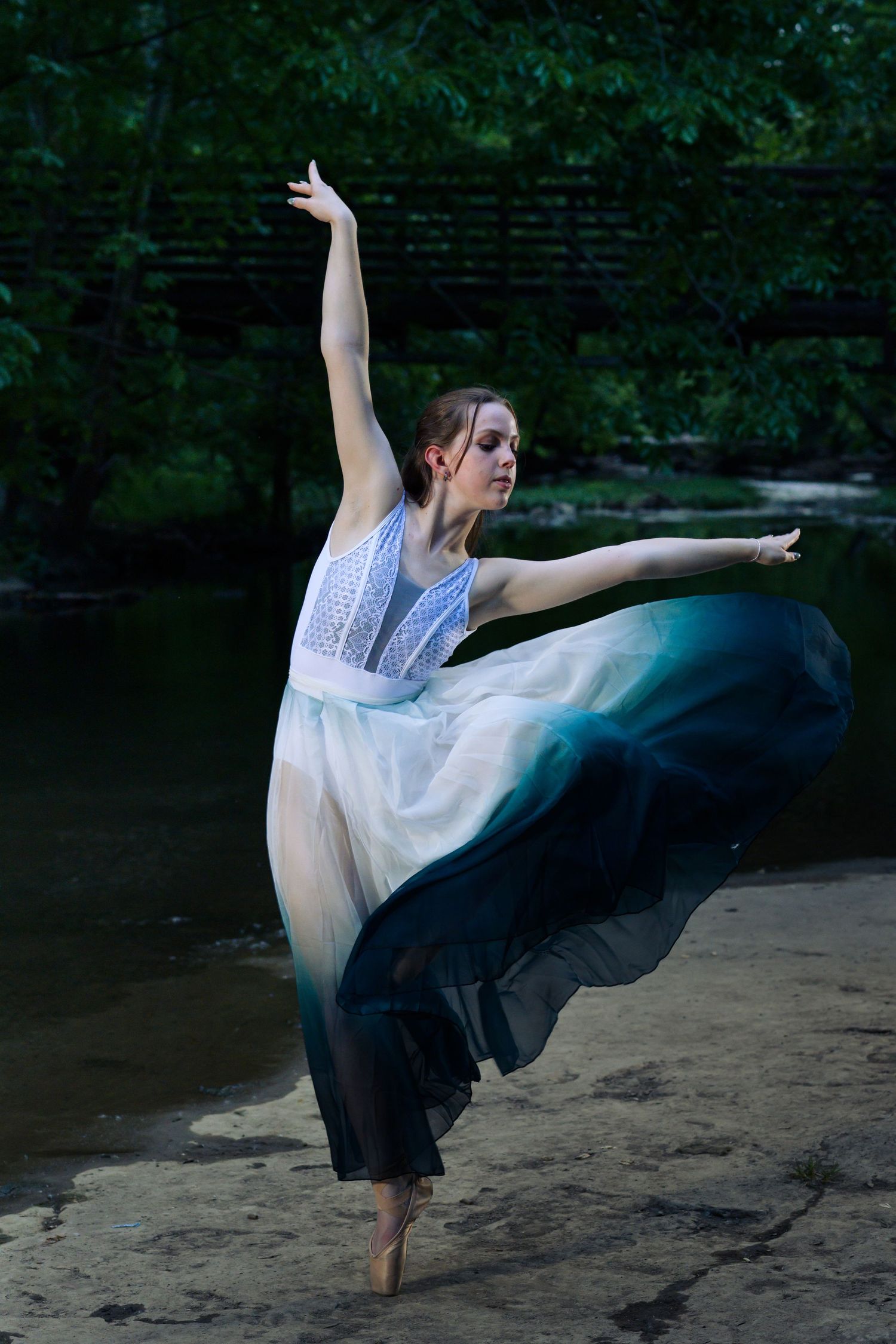 A graceful ballerina in a flowing white and teal ombre dress poses on one foot outdoors near a serene forest stream.