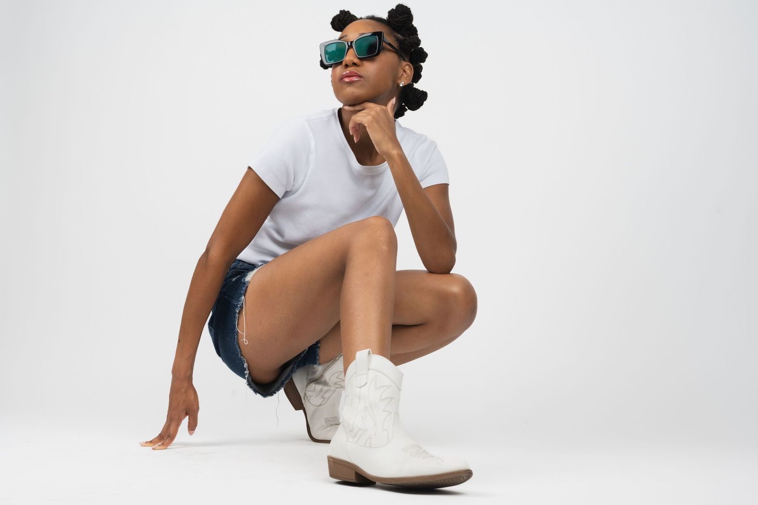 A model in a white t-shirt, denim shorts and white boots crouches while wearing green sunglasses against a white backdrop.