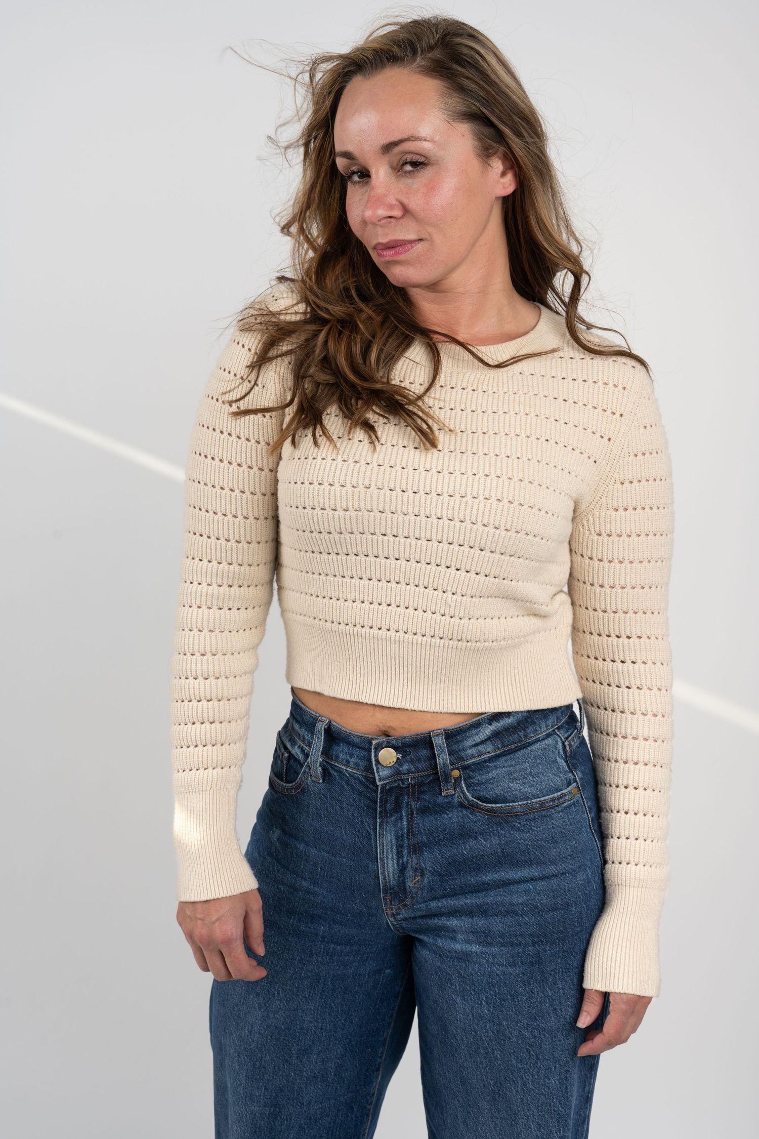 Woman in cream knit crop top and jeans poses in studio setting.