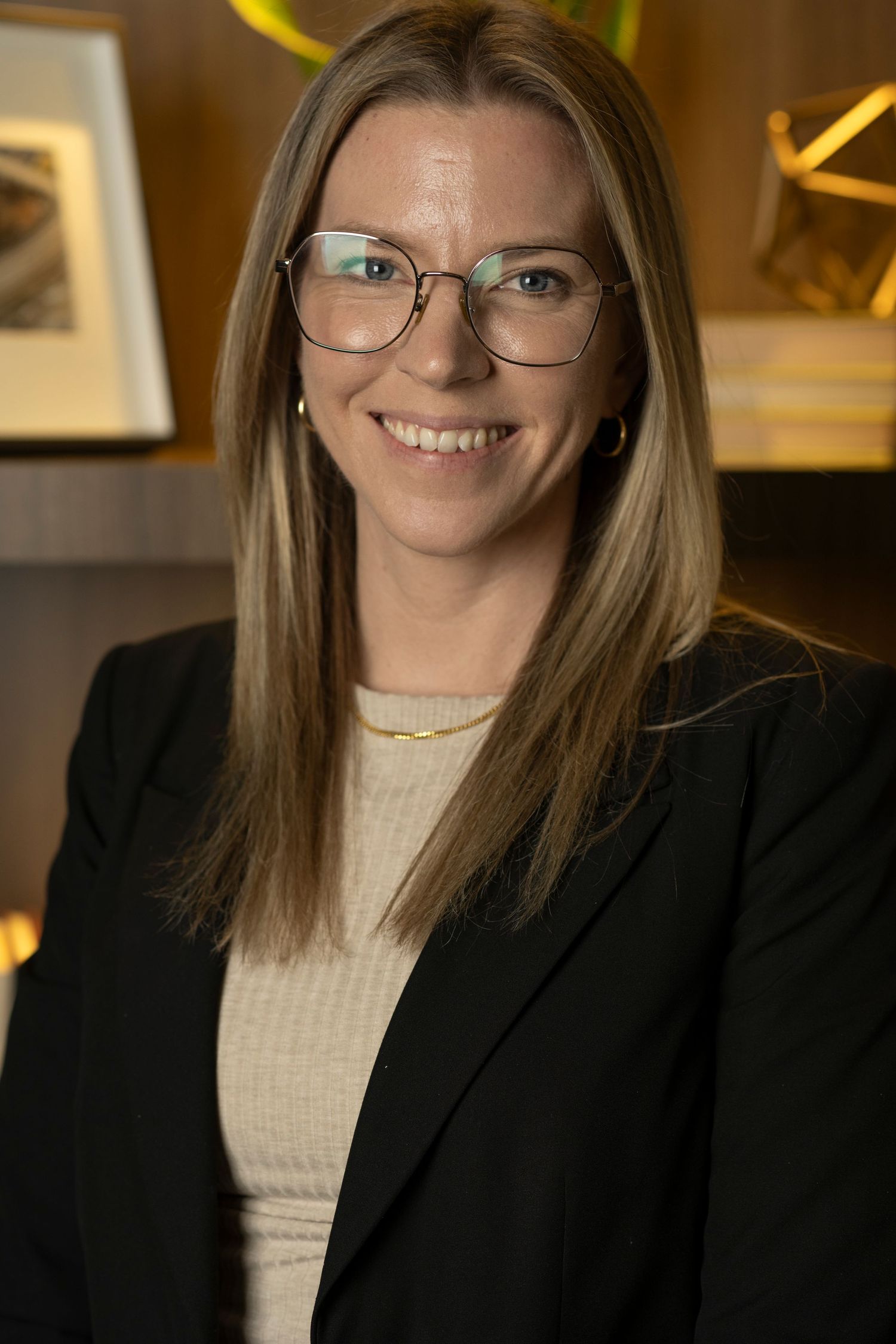 Professional headshot of person in black blazer and cream sweater with glasses in warm-lit setting.