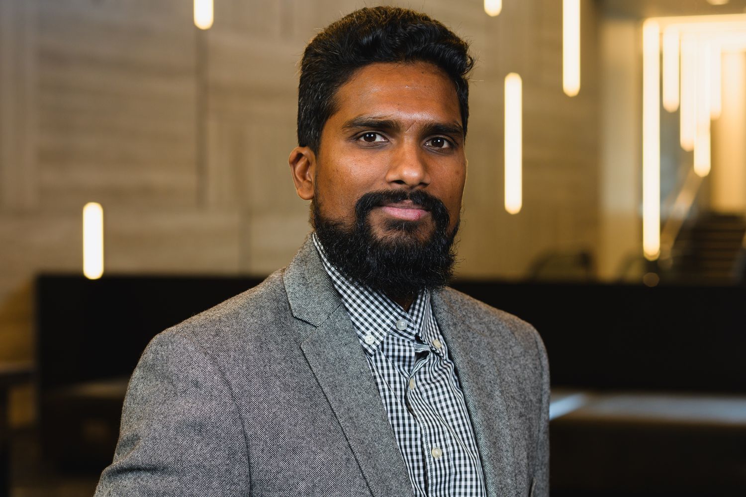 Business headshot of person in checkered shirt and grey blazer against office backdrop.