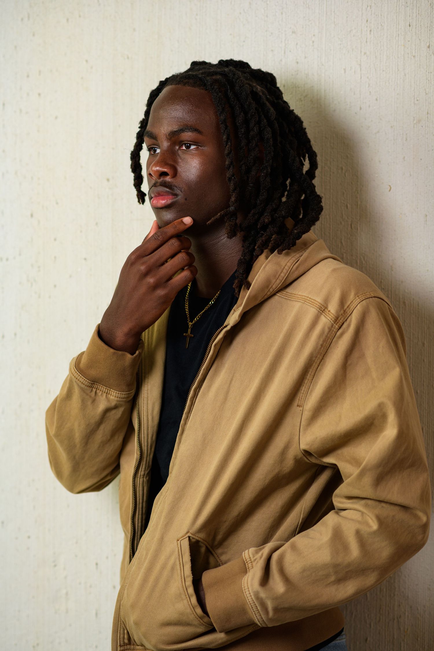 Young Black man with dreadlocks in tan jacket, hand on chin, thoughtful pose against neutral wall.