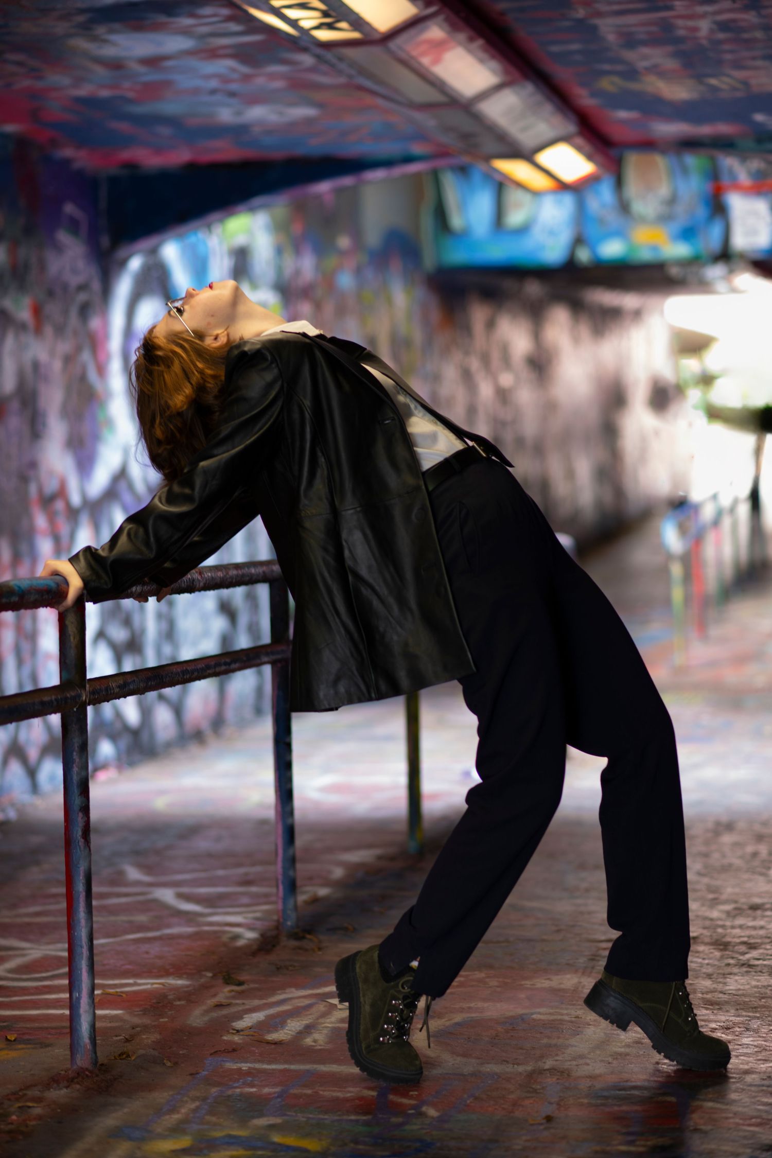 A person in black clothing leans back dramatically against a railing in a graffiti-covered tunnel.