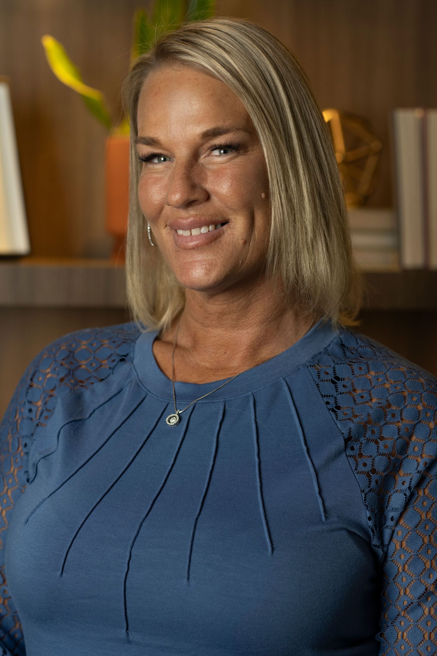 Business portrait of person wearing blue lace-detailed top in warm-lit office setting.