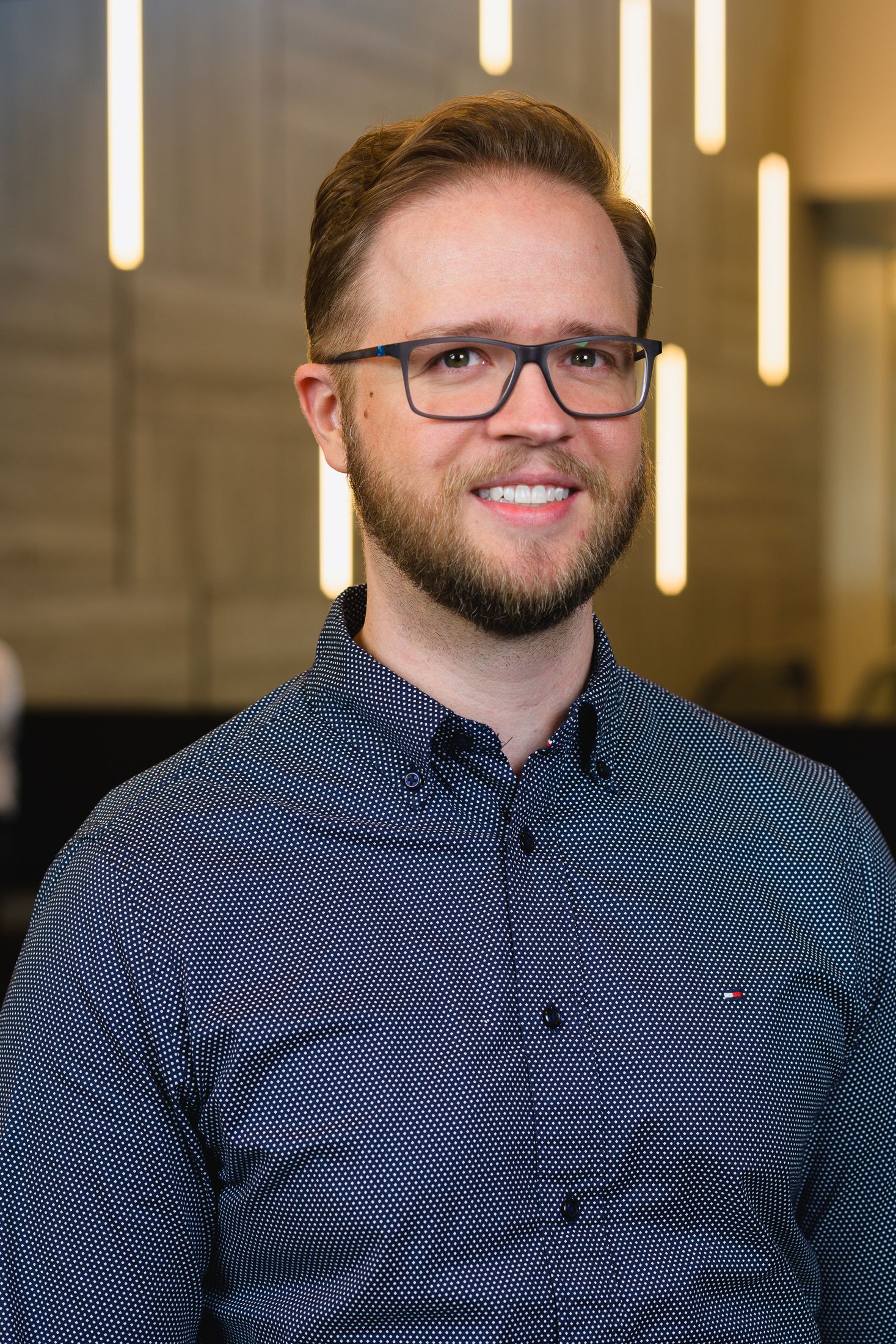 Professional headshot of person in navy blue button-down shirt with glasses against office background.