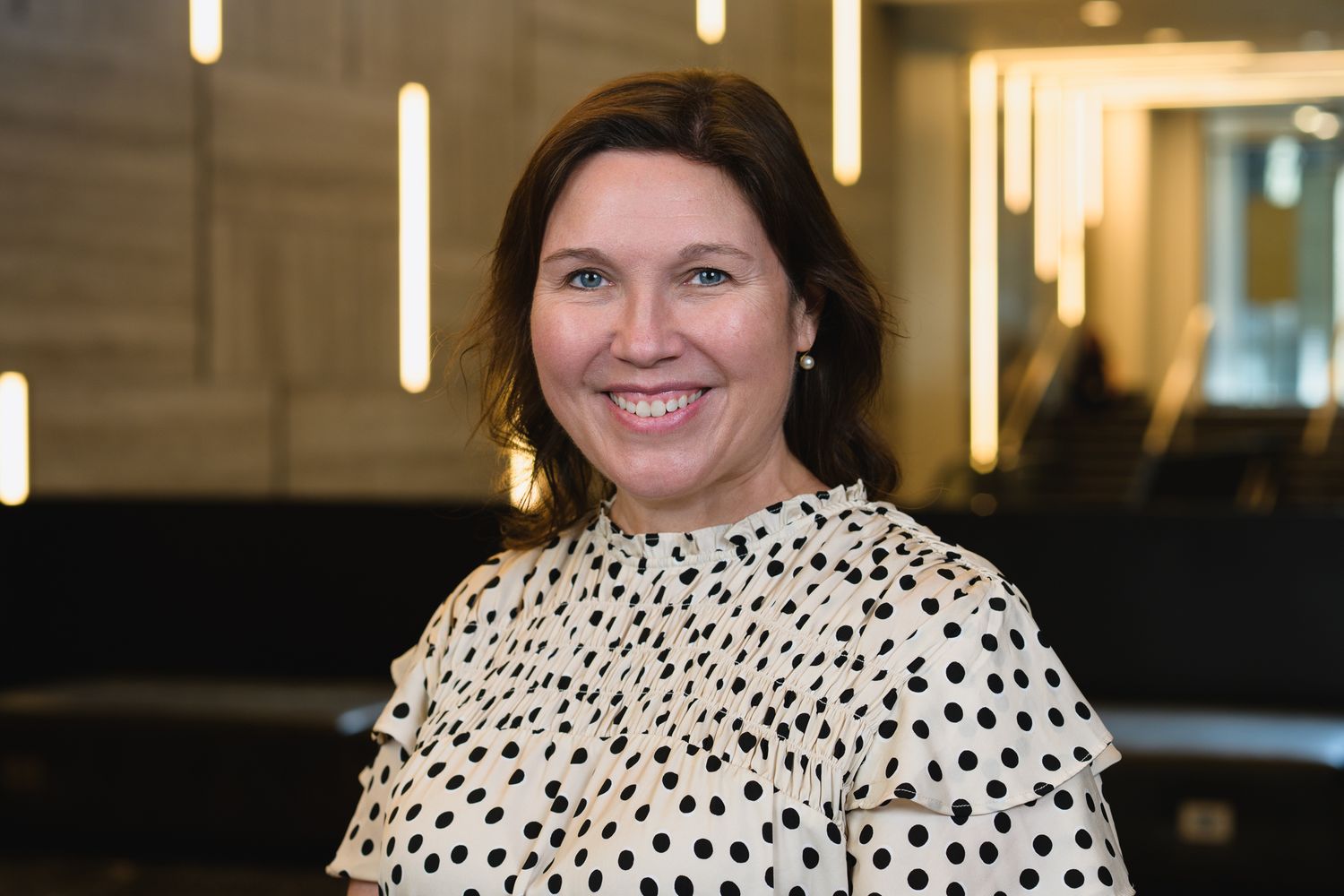 Professional headshot of person in polka dot blouse smiling against office backdrop.