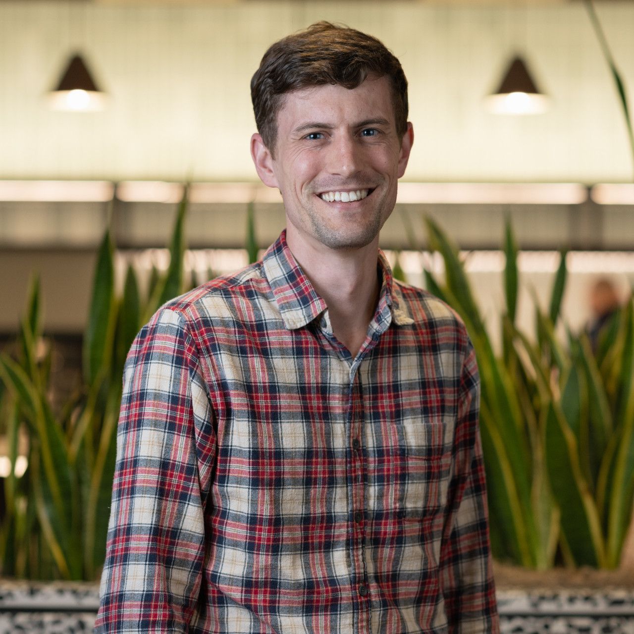 Professional portrait in plaid button-up shirt with office plants in background.