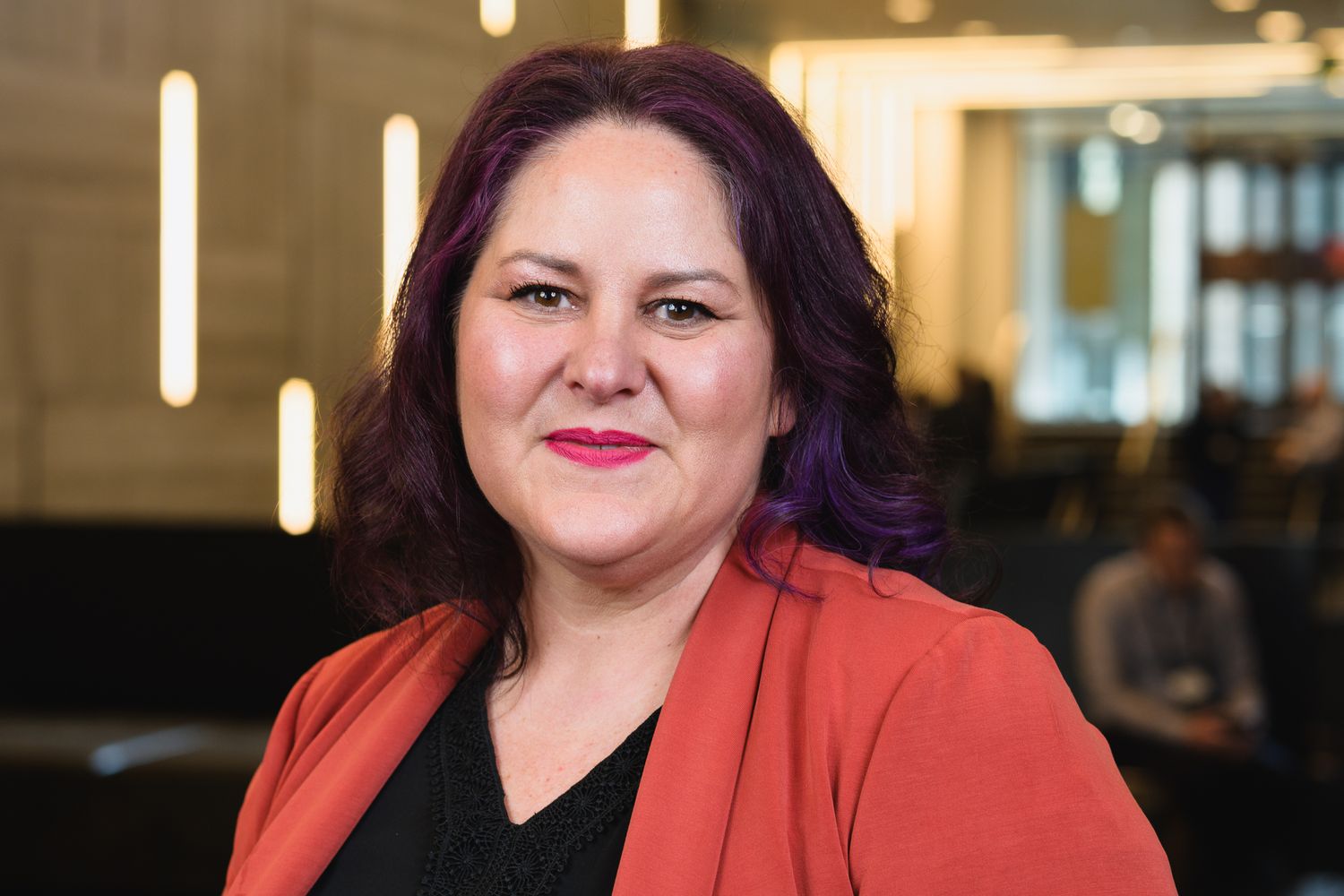 Professional portrait of person in red blazer against modern office interior.