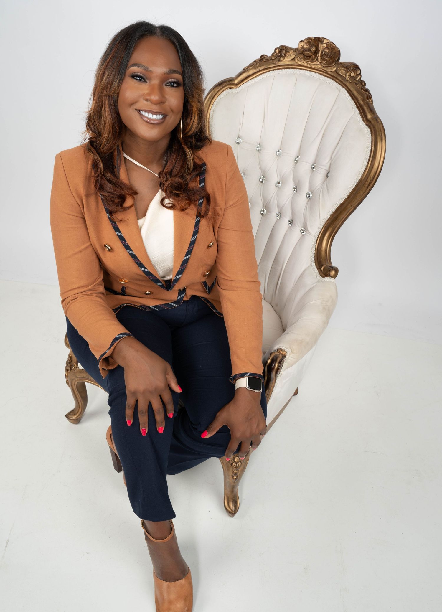 Professional woman in camel blazer and dark jeans poses confidently on elegant white and gold chair.