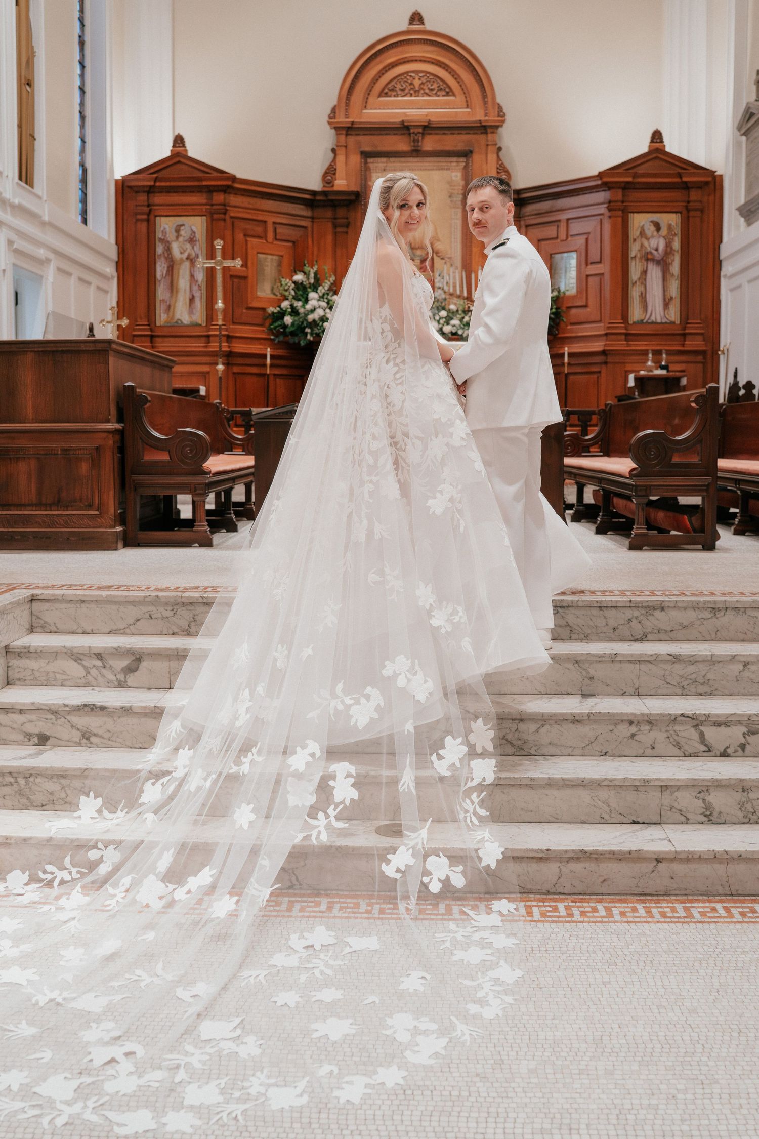 Bride and groom pose at church altar, bride wearing stunning lace gown with long floral cathedral veil trailing on marble floor.