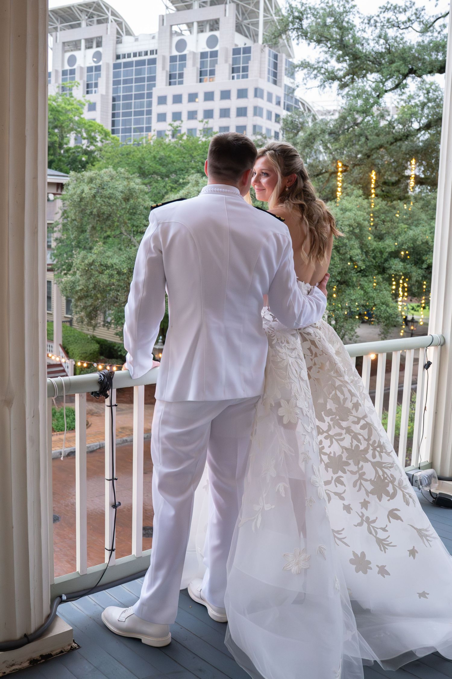 A bride and groom in white wedding attire stand on a balcony overlooking lush greenery and city buildings.