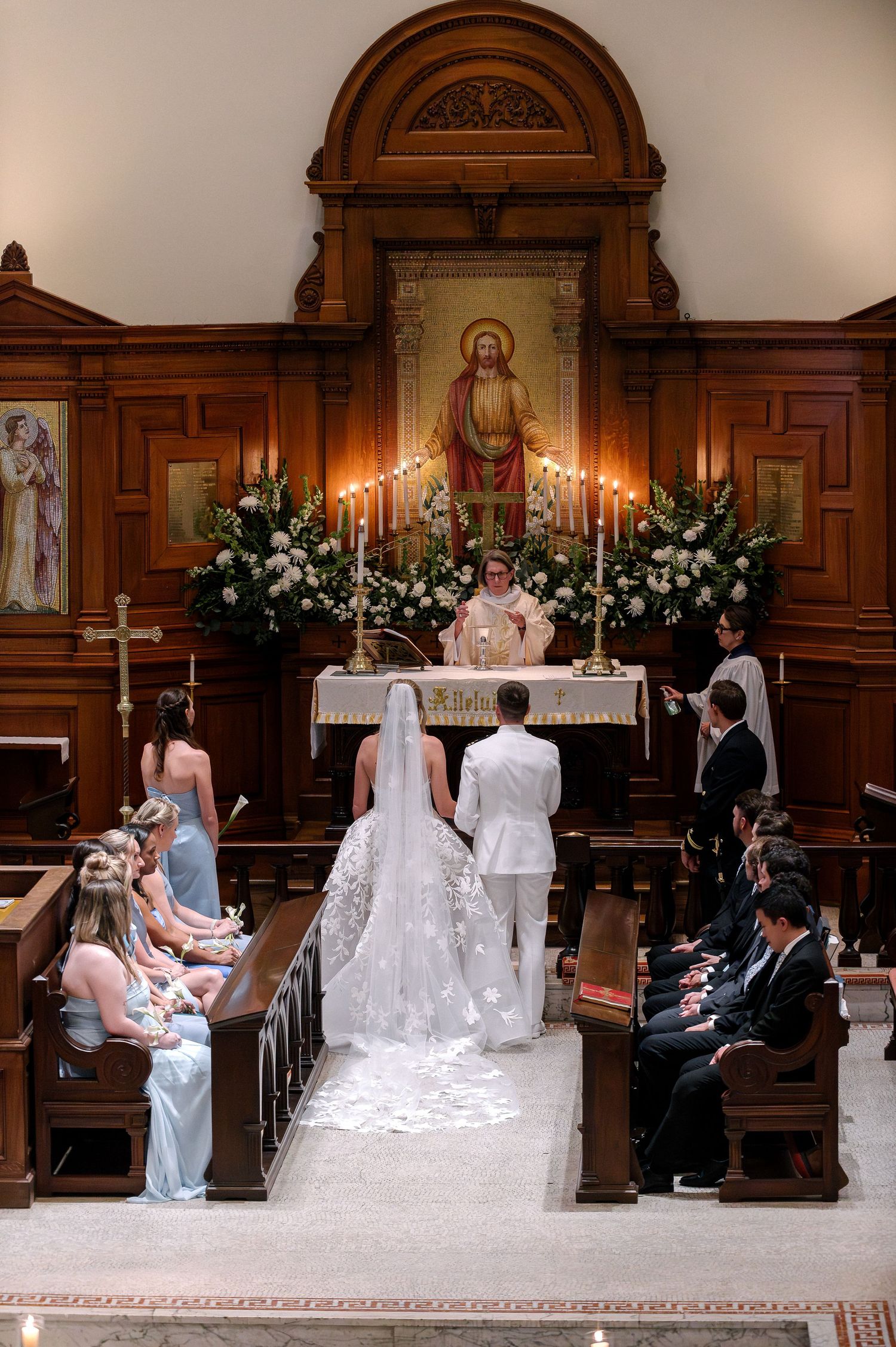 Bride and groom stand at church altar during wedding ceremony with bridesmaids seated in pews and floral arrangements.