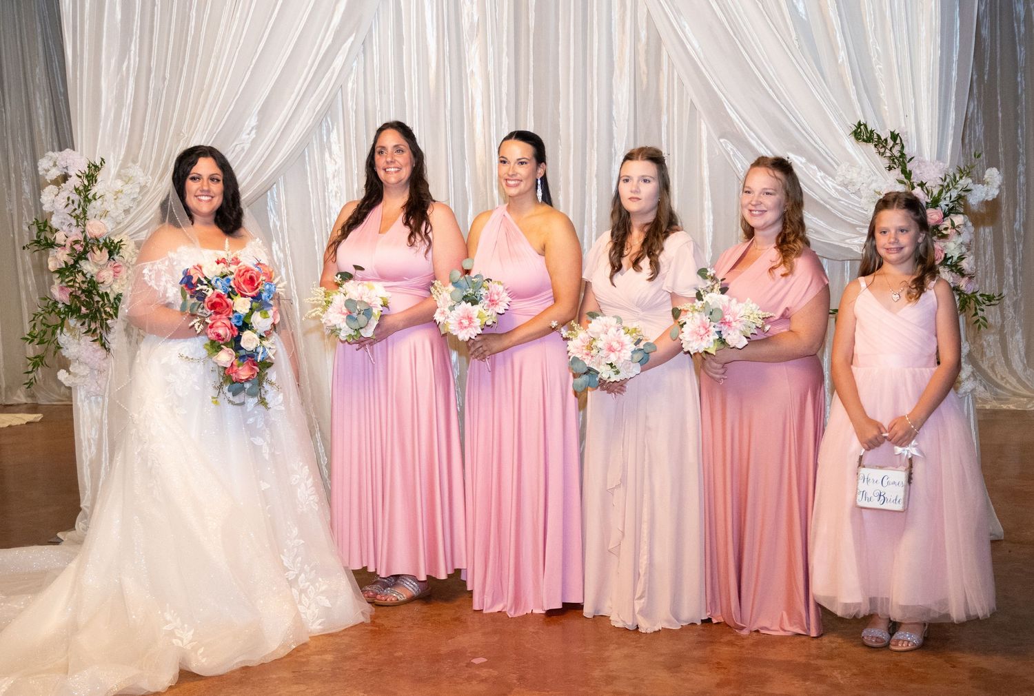 Bridal party in pink dresses poses against white draped backdrop with floral decor.