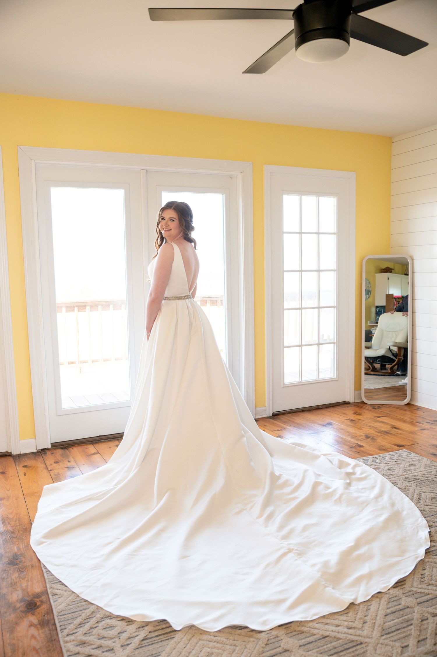 Elegant white wedding gown with long train displayed in sunlit room with yellow walls.