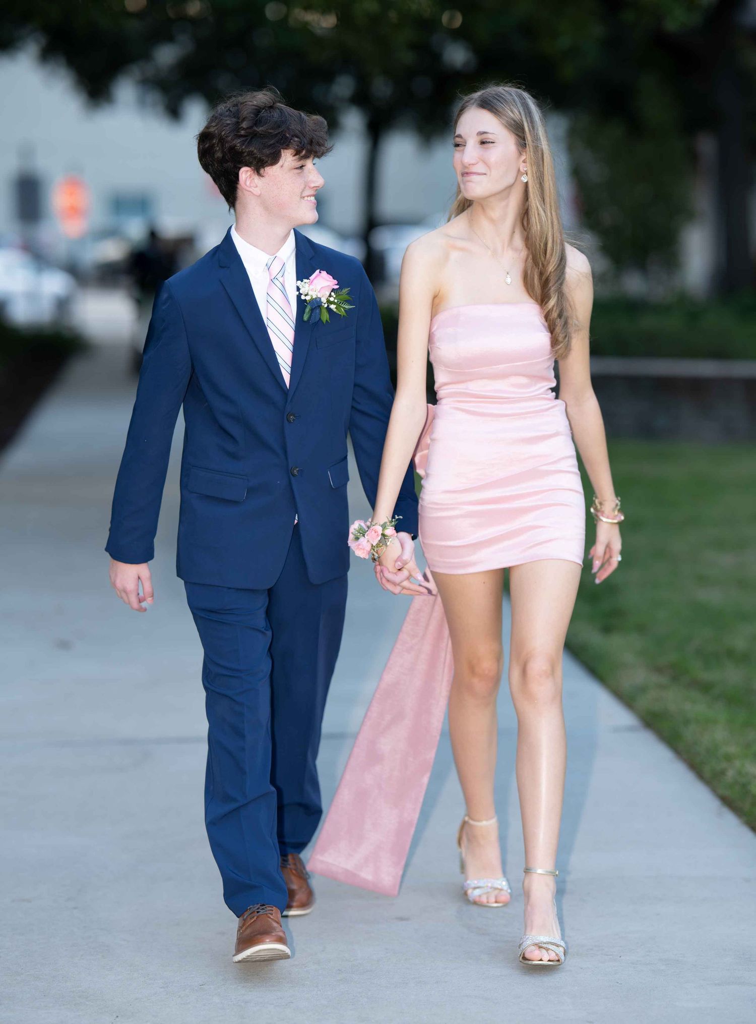 A couple dressed in formal attire, with a navy suit and pink dress, walking hand in hand on a sidewalk at dusk.