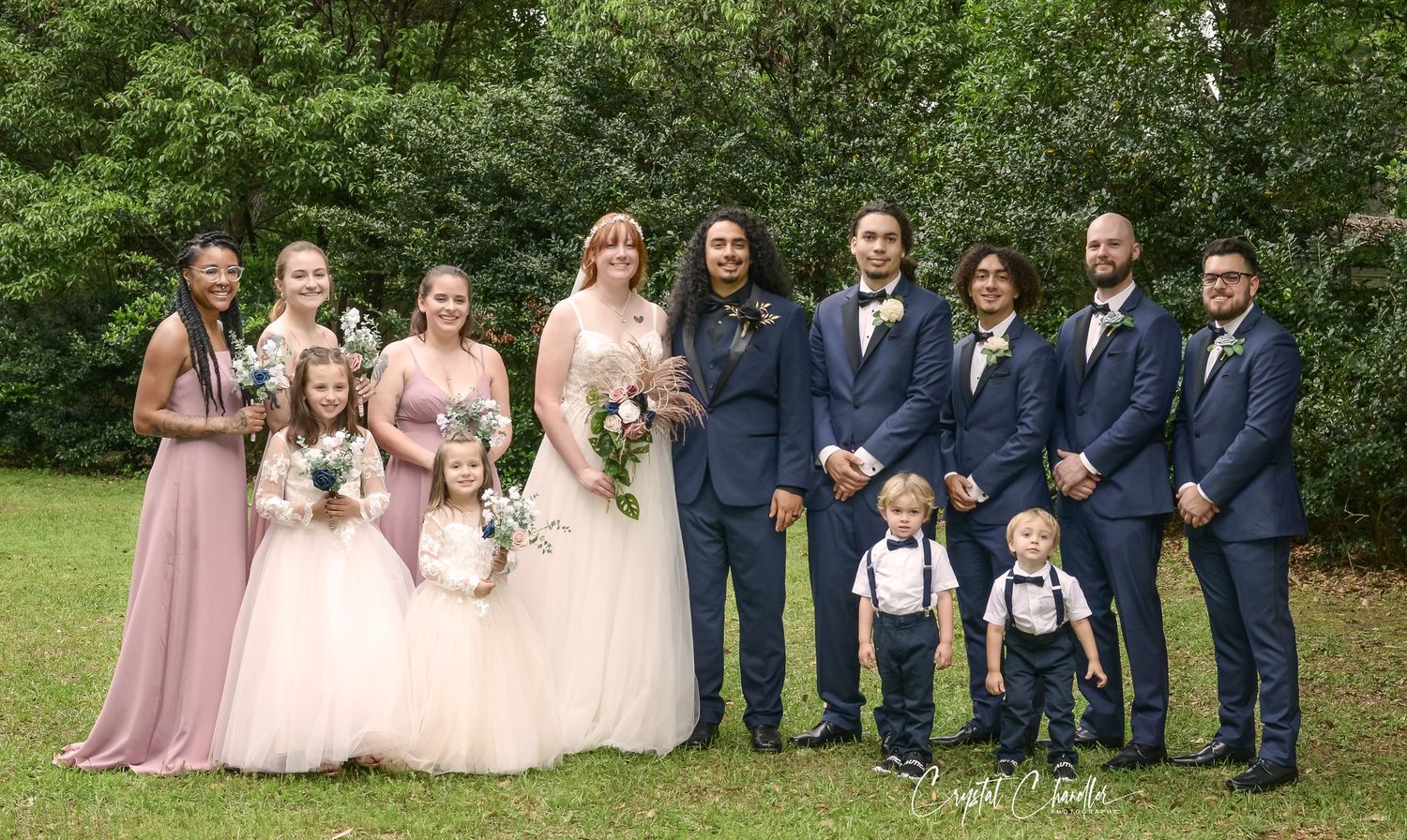 A wedding party poses outdoors in navy suits and pink dresses against a green natural background.