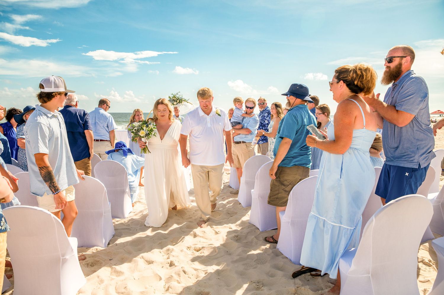 Beach wedding celebration with guests in light colored attire gathered on sandy shore.