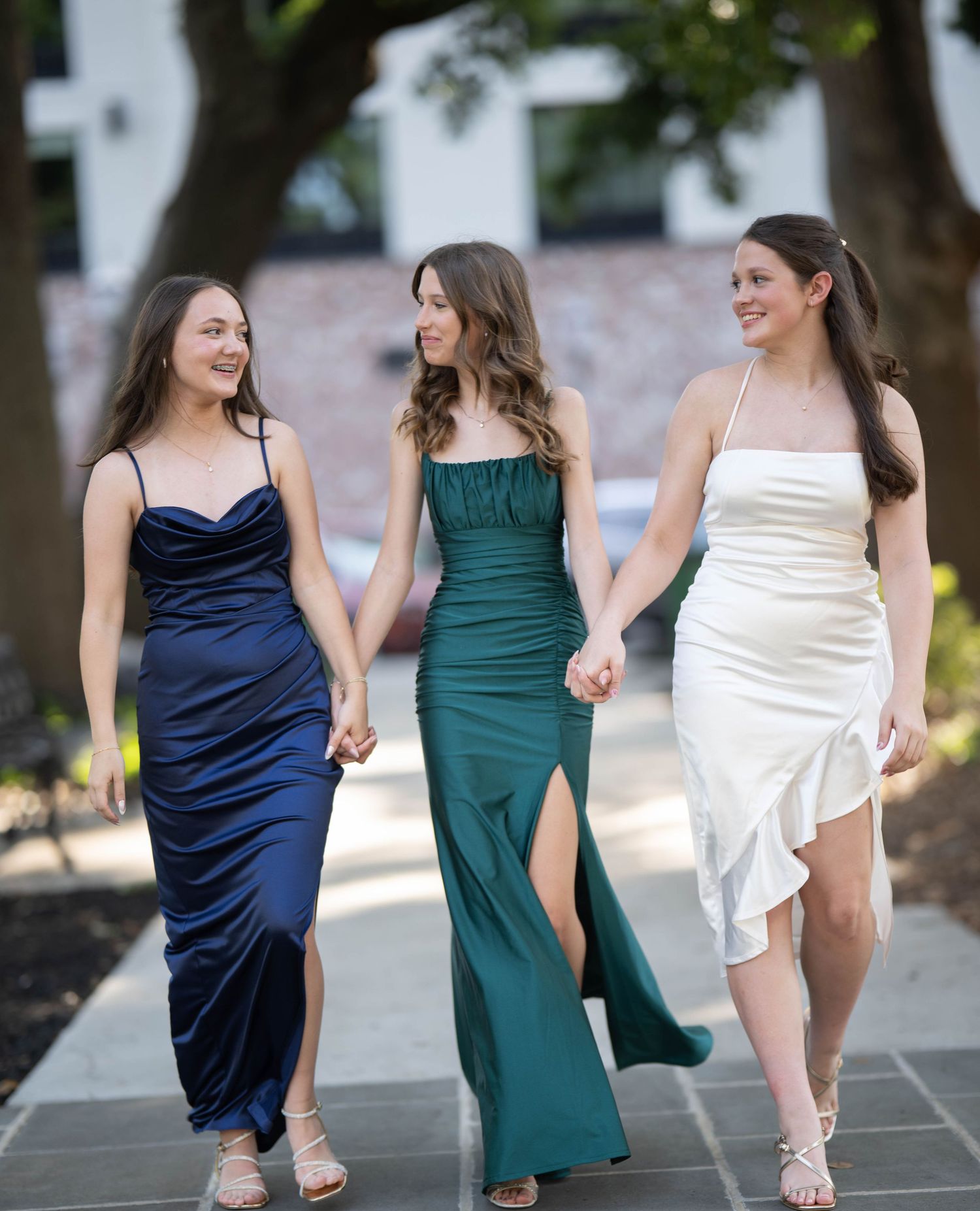 Three friends in elegant evening gowns - navy blue, emerald green, and white - walk together outdoors while holding hands.