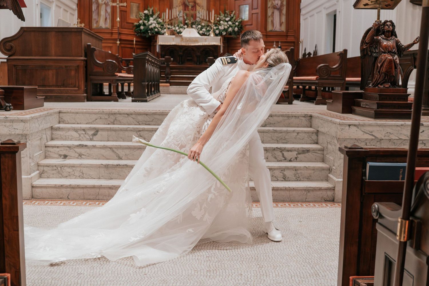 Bride and groom share a romantic dip kiss inside an elegant church with grand staircase and wooden pews.