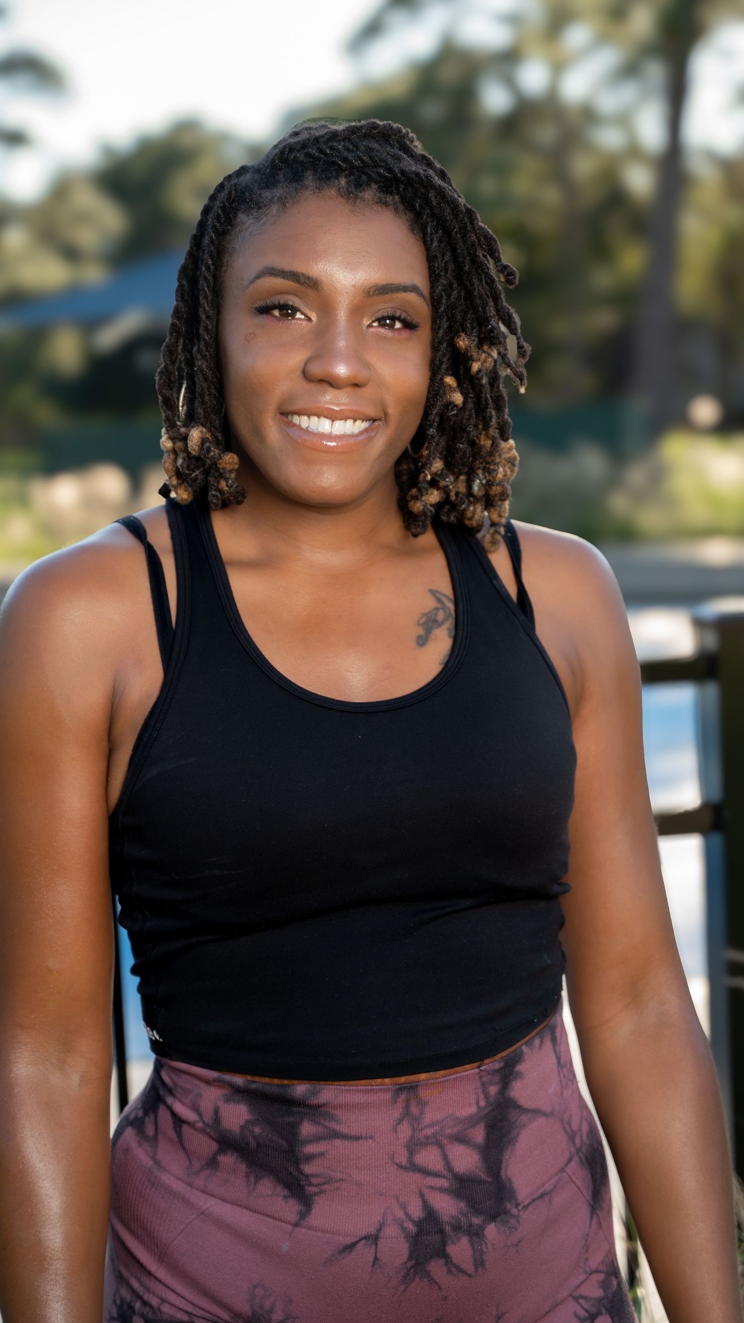 A person wearing a black sleeveless top and purple tie-dye bottoms poses outdoors with a bright smile.