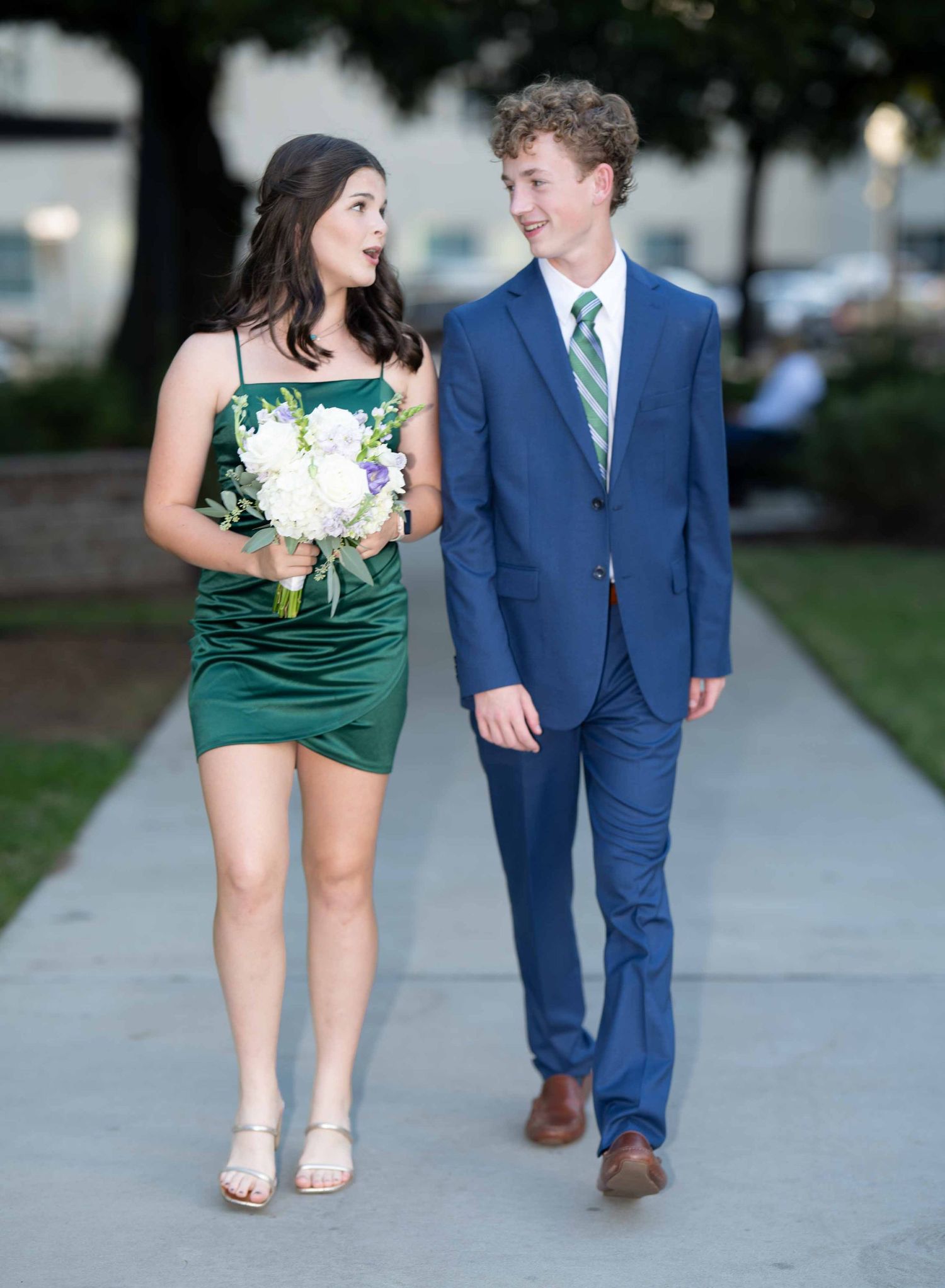 A couple in formal attire walking together on a sidewalk, with the woman in green and the man in a blue suit.