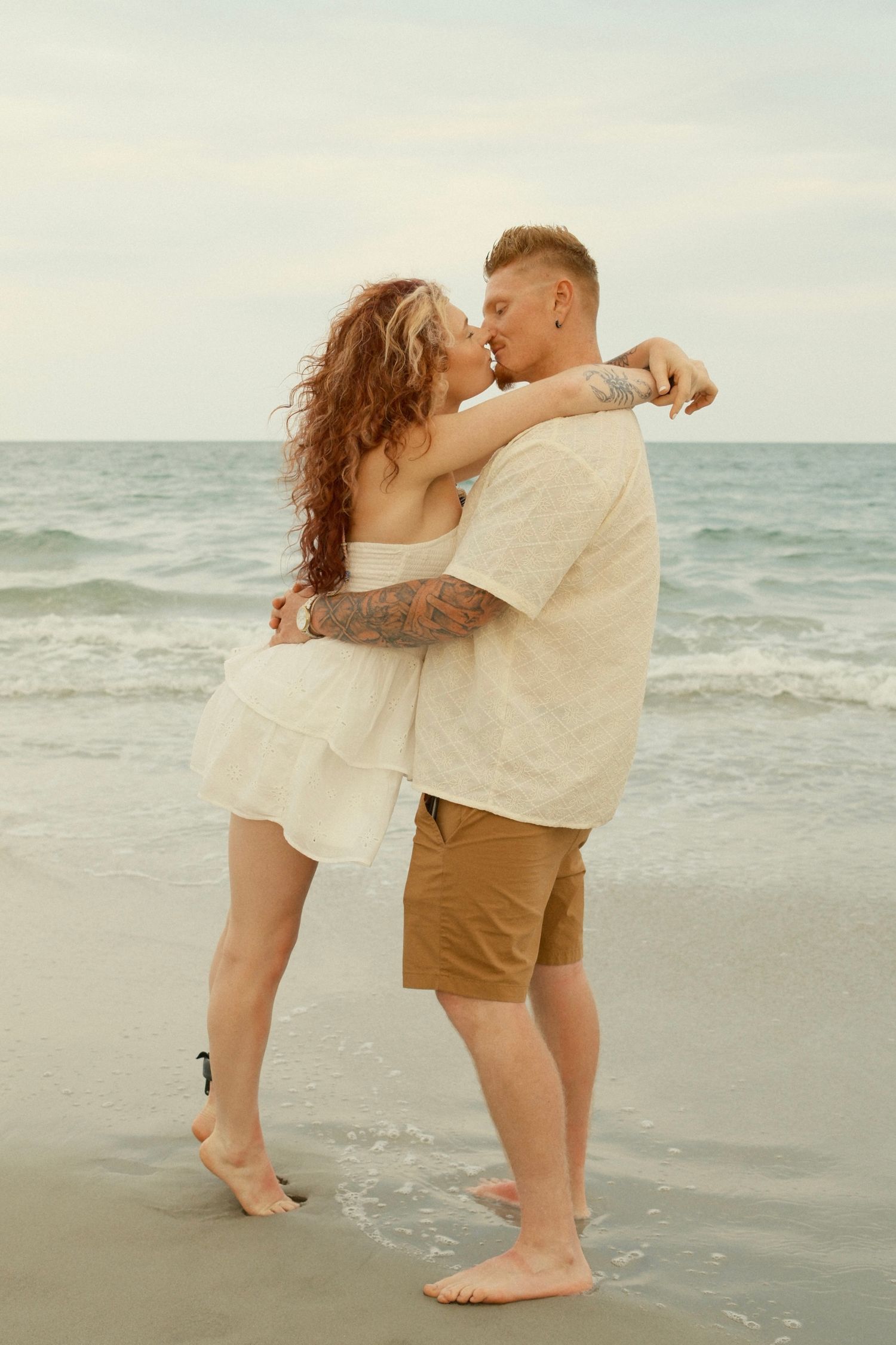 A romantic couple embraces and kisses on a serene beach during sunset with ocean waves in the background.