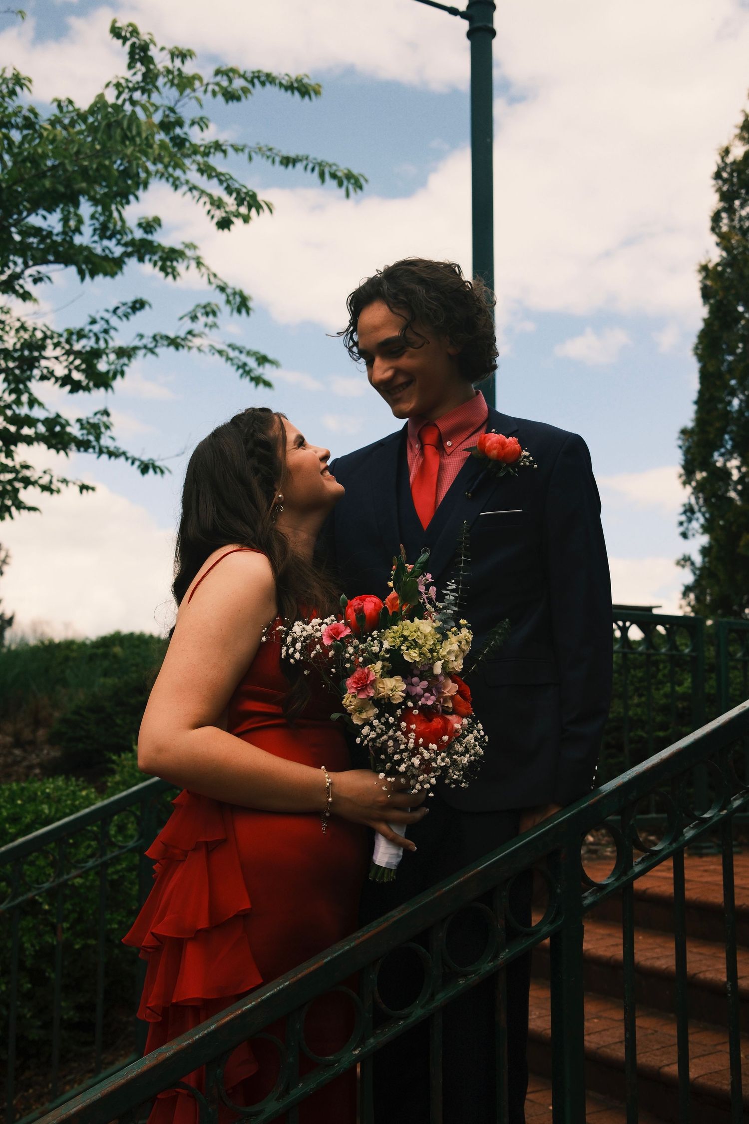 A couple in formal attire share romantic moments on outdoor stairs, with the person in red wearing a strapless dress.