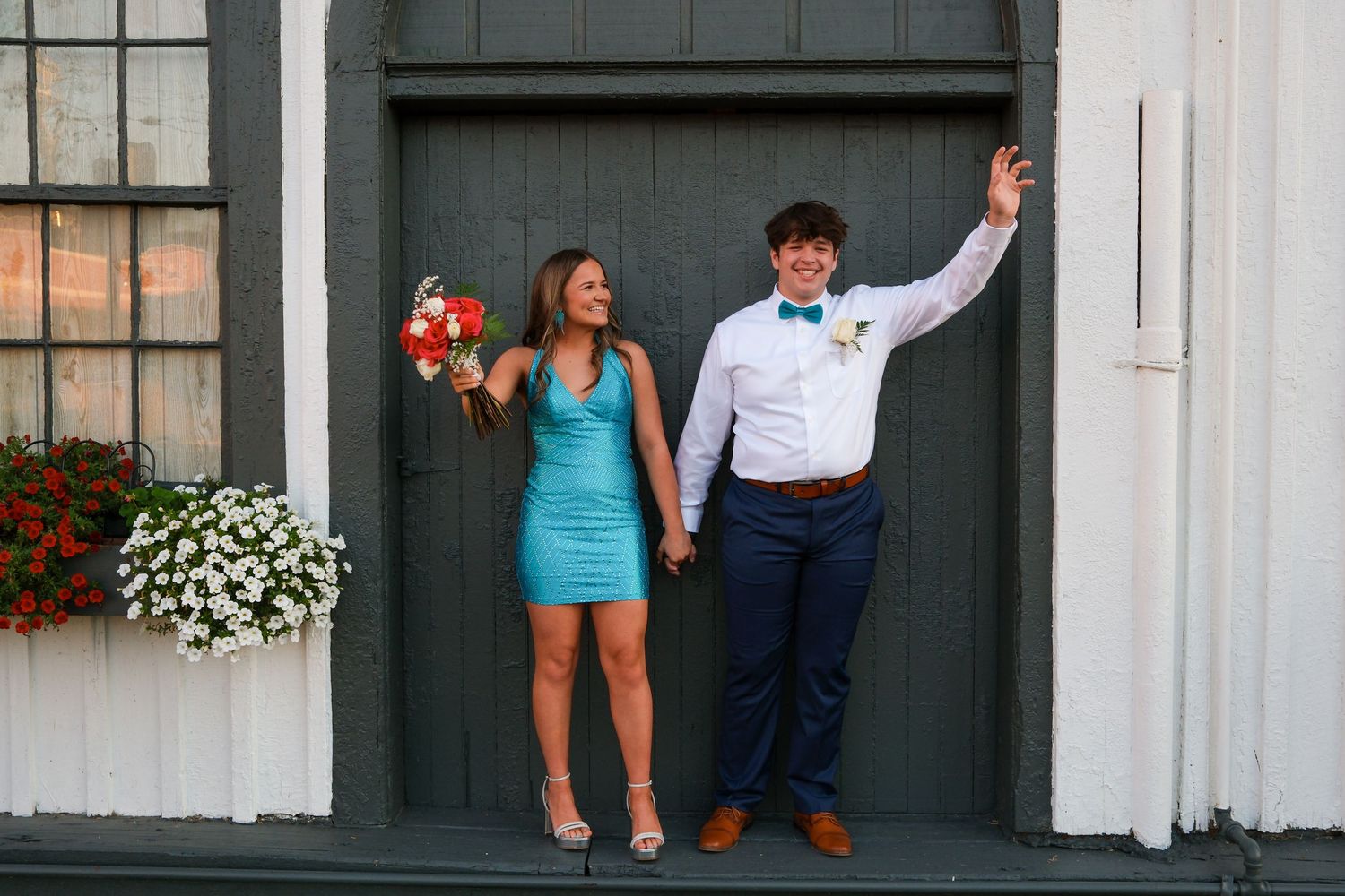 Couple in formal attire strikes fun pose against dark gray door with white building and flowers.