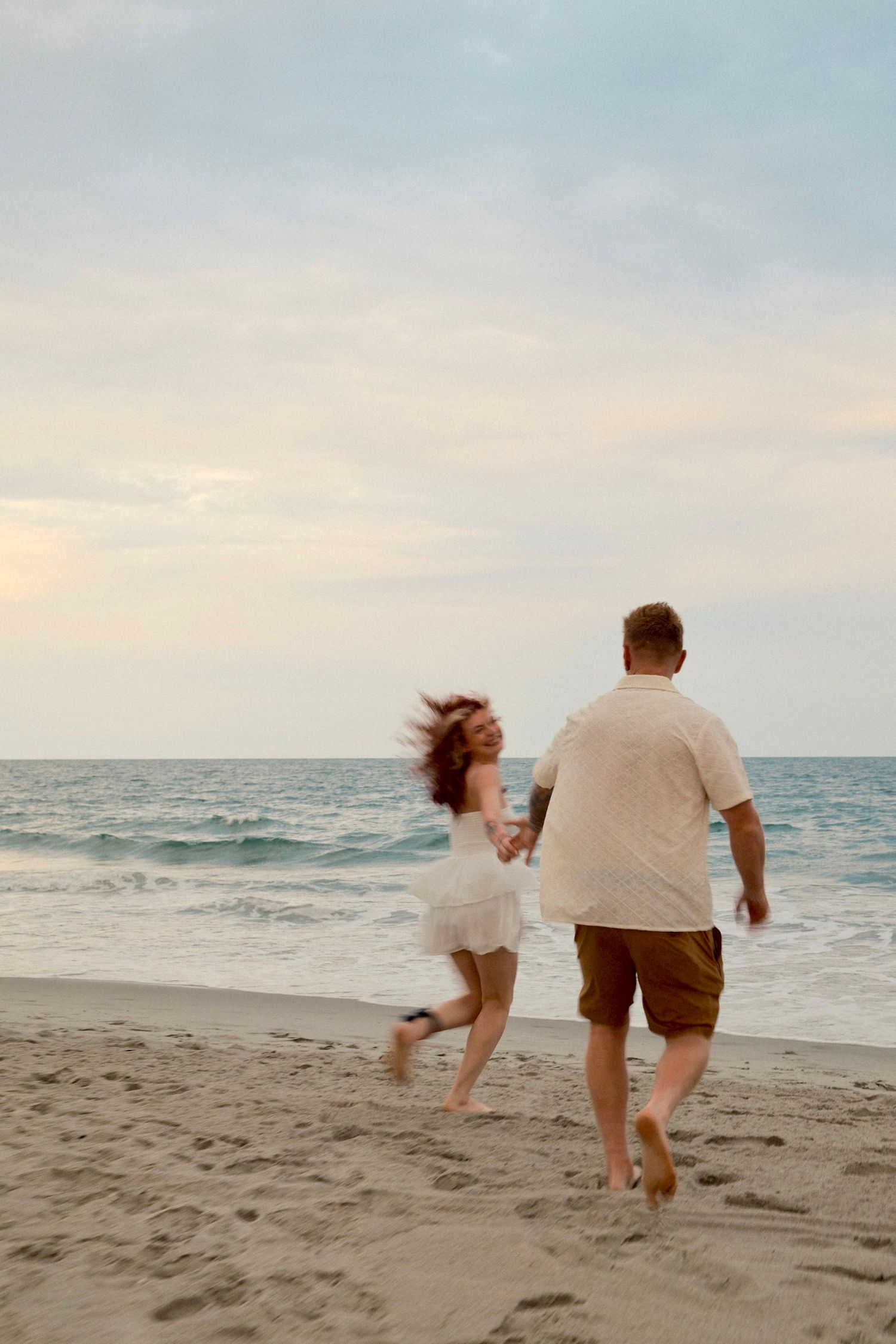 Two people run playfully along a sandy beach at sunset with ocean waves in the background.