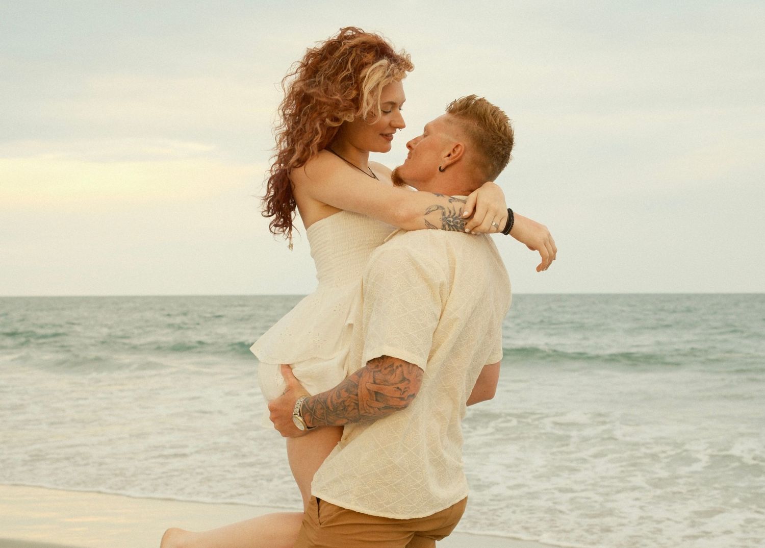 Couple embracing while standing on sandy shoreline with ocean waves in background.