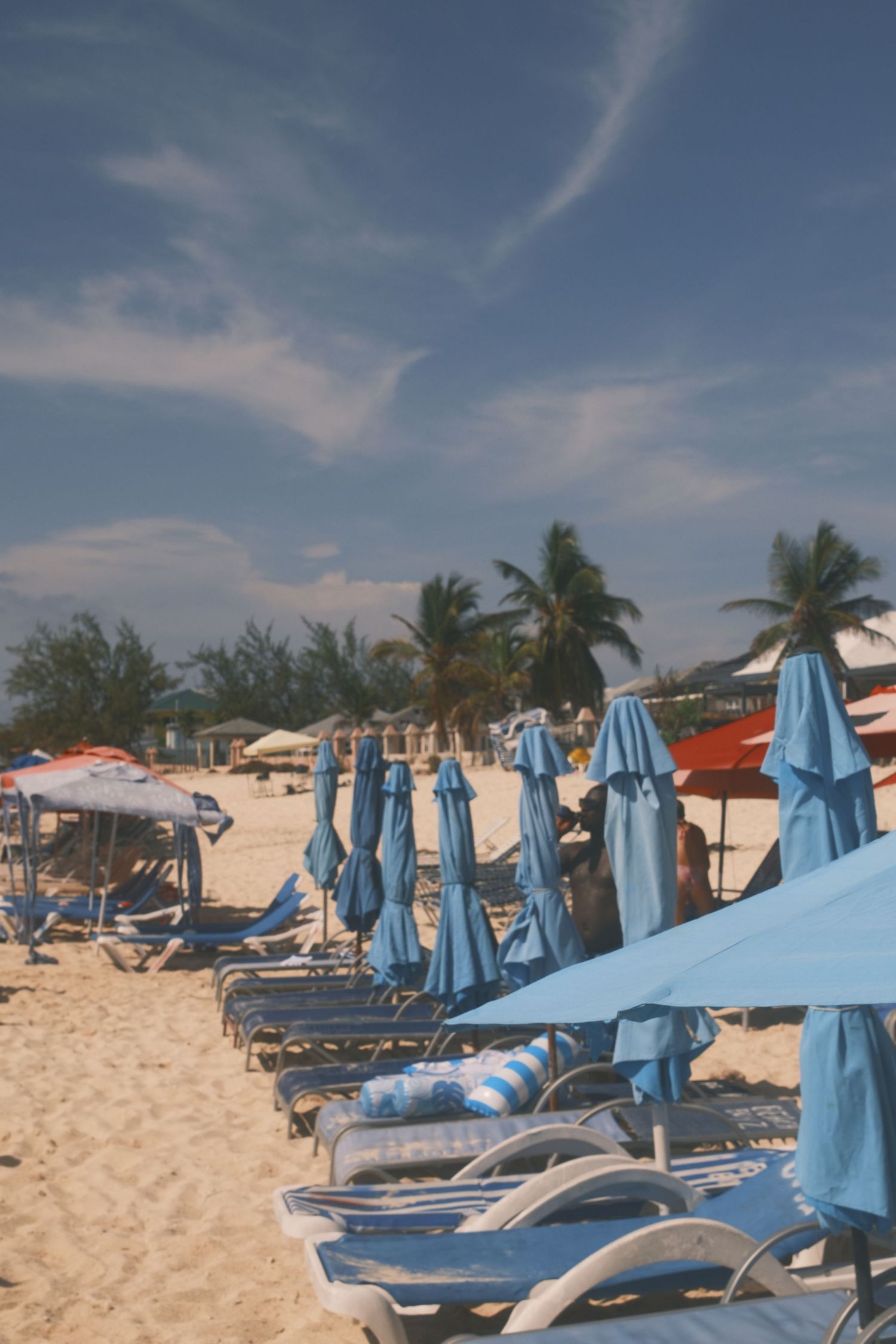 Beach lounge chairs with blue umbrellas line a sandy tropical shore with palm trees and clear skies overhead.