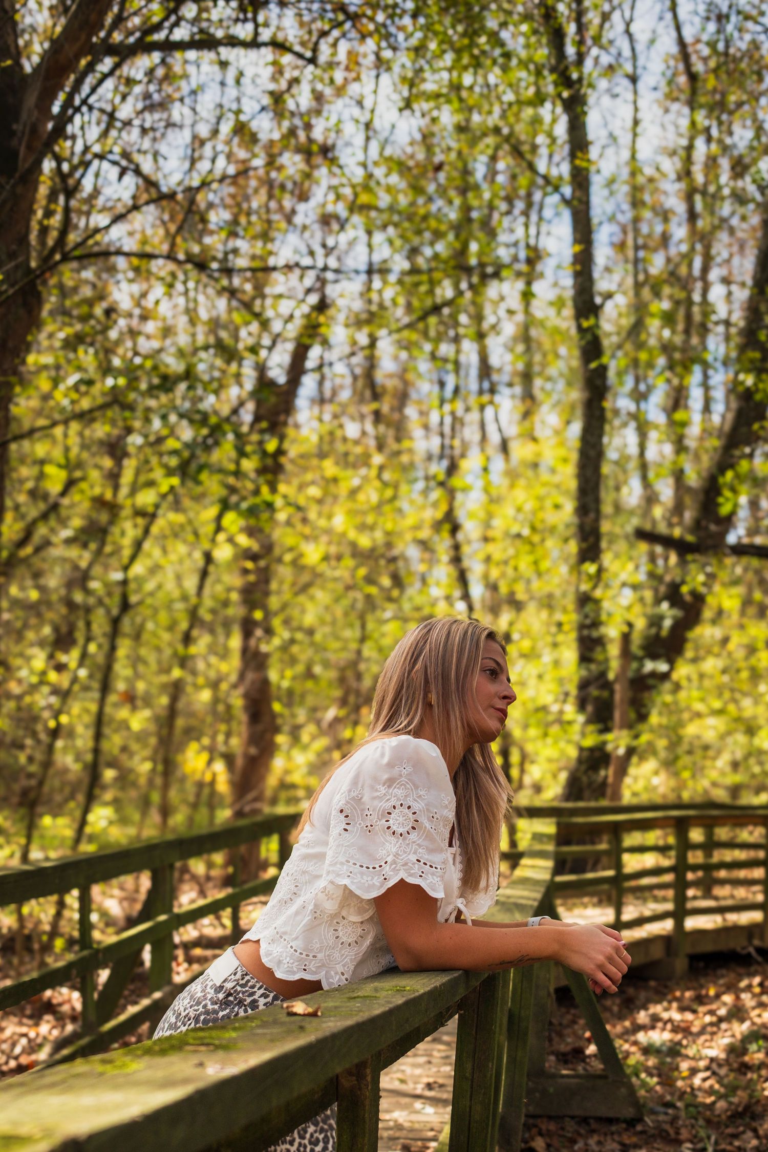 Person in white lace dress leans on wooden railing in sunlit forest with autumn foliage.