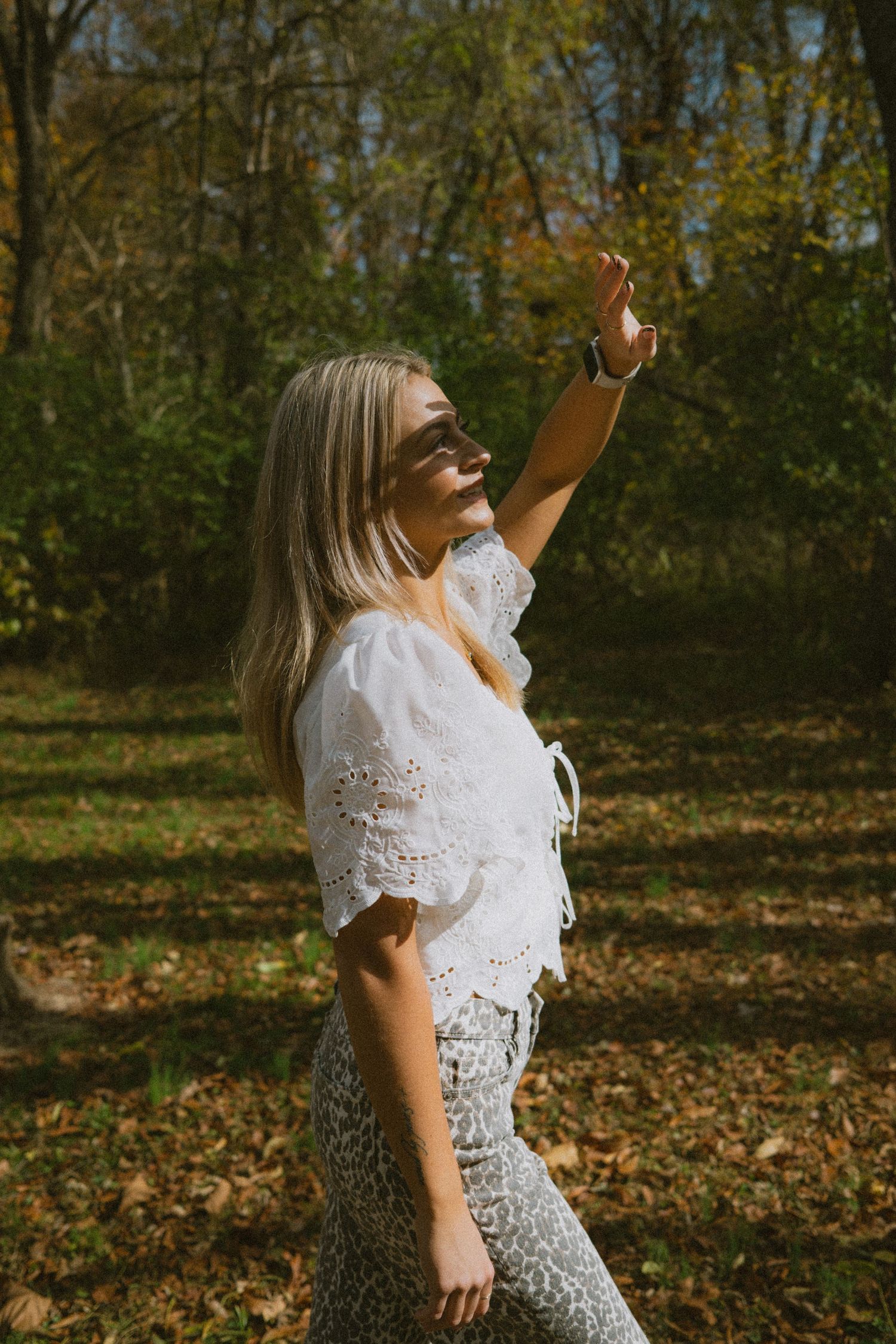 A person in a white lace top and patterned pants reaches upward while standing in a wooded area with autumn leaves.
