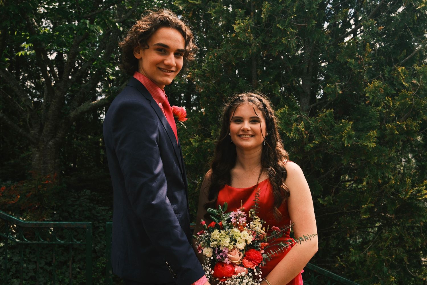 A couple in formal attire poses outdoors with colorful bouquets against a natural green background.