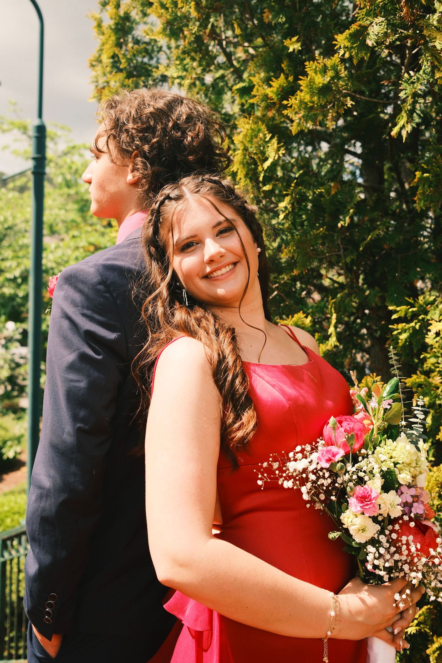 People in red dresses gather outdoors with floral bouquets under trees on a sunny day.