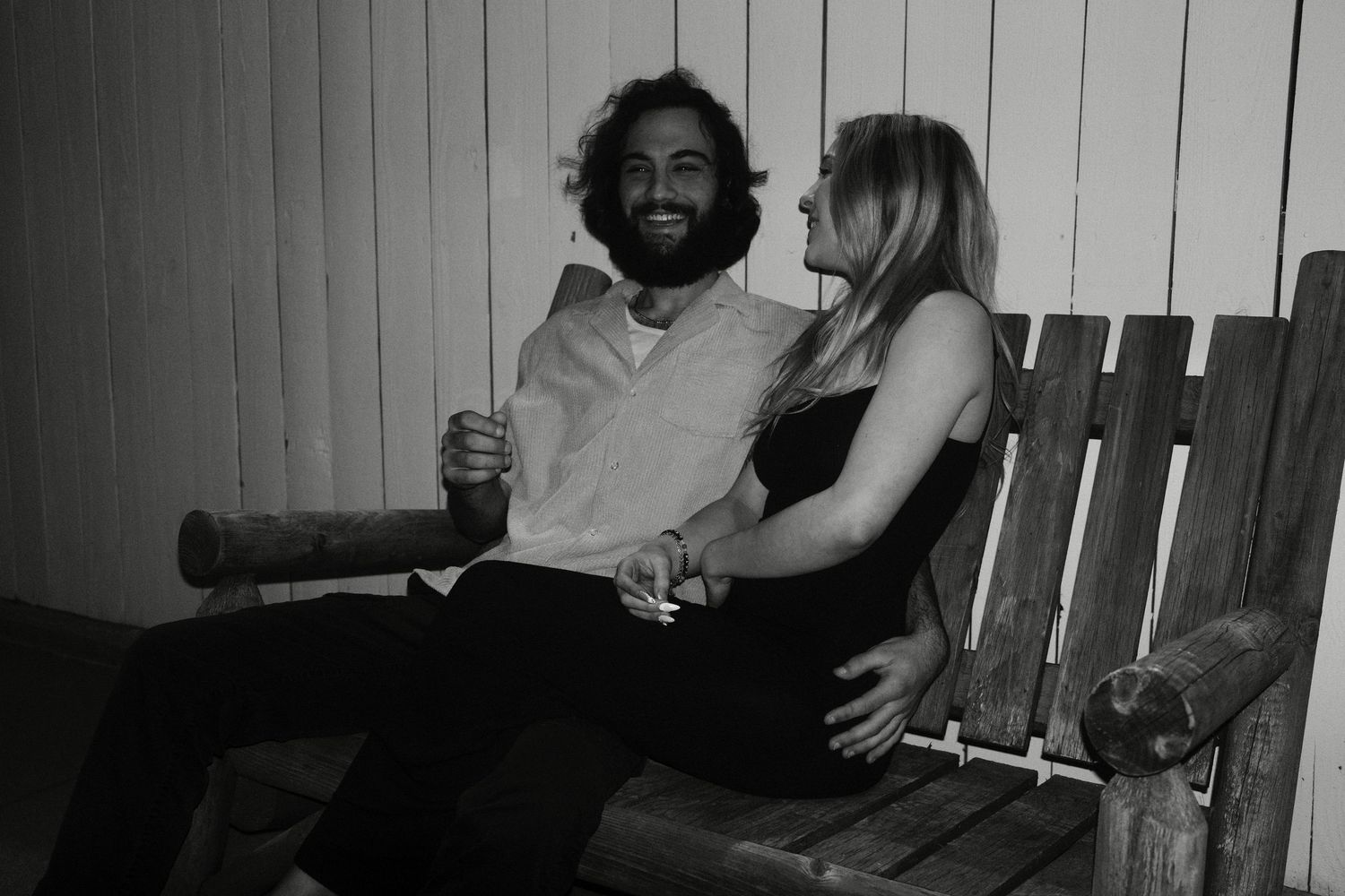 A couple sits intimately together on a wooden bench in a black and white photograph.