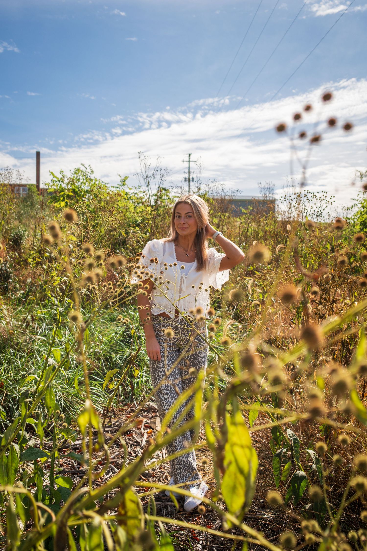 A person stands in a sunlit field of wildflowers and tall grass against a bright blue sky with power lines in the distance.