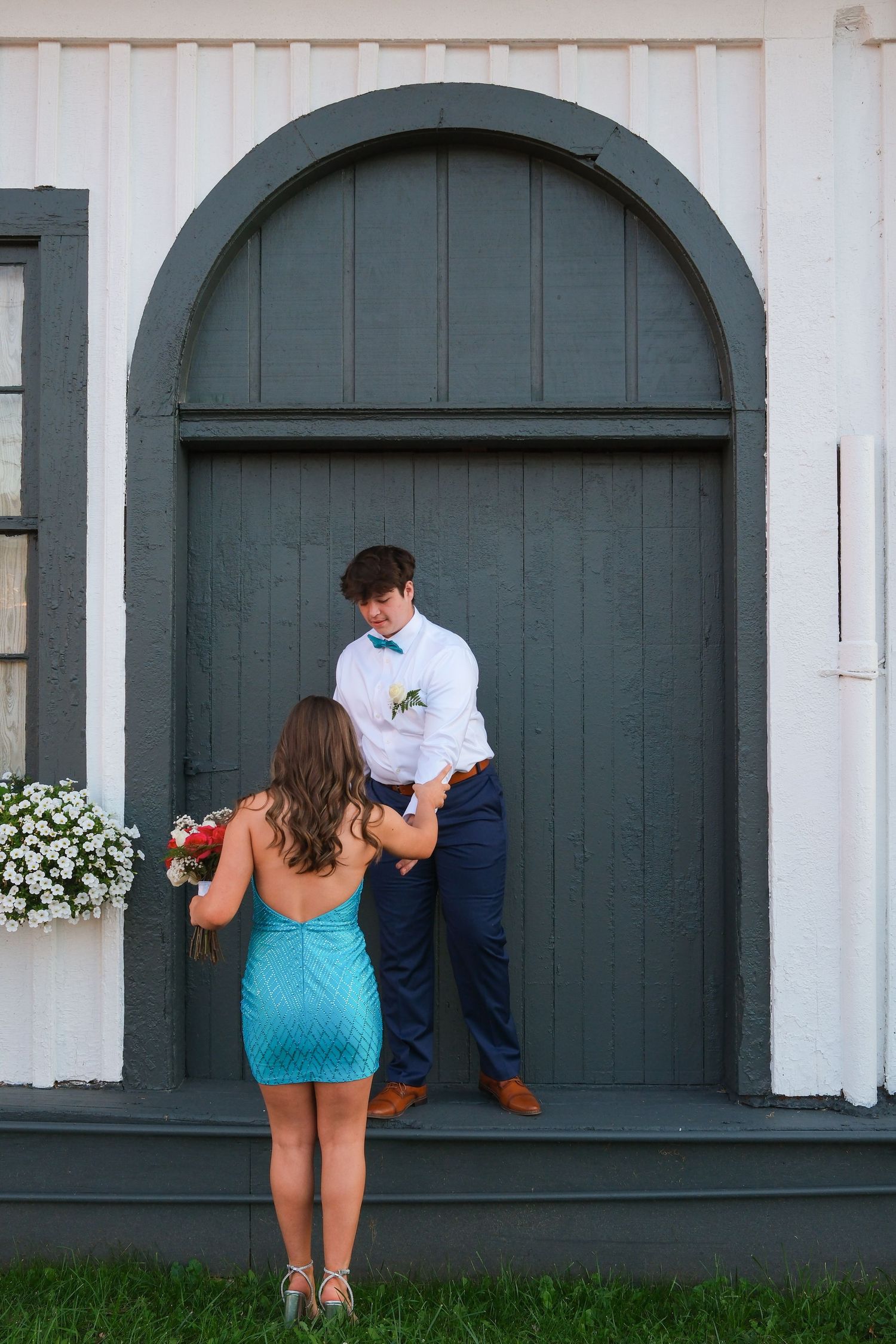Couple poses in front of a large arched gray door with white flowers on the side of a building.