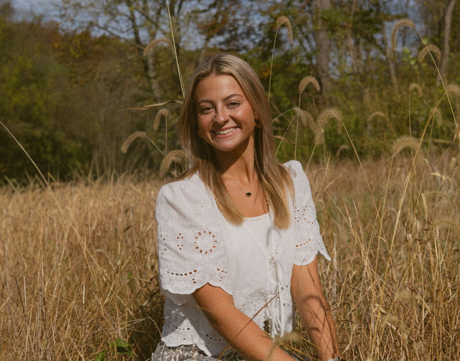 A person in a white eyelet dress sits in a golden field of tall grass during autumn.