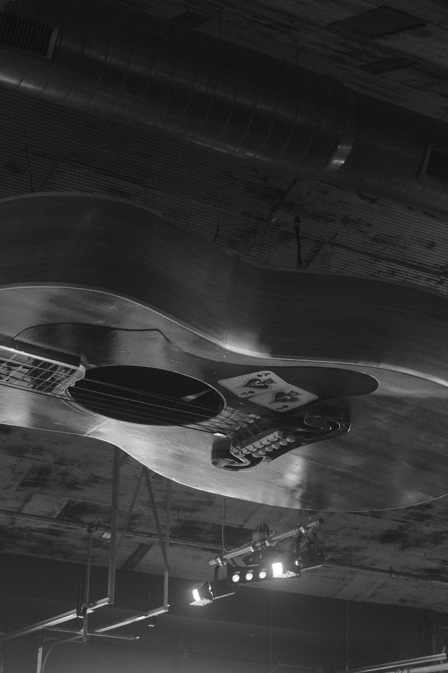Black and white photograph of an acoustic guitar mounted on a dark ceiling with stage lighting visible below.