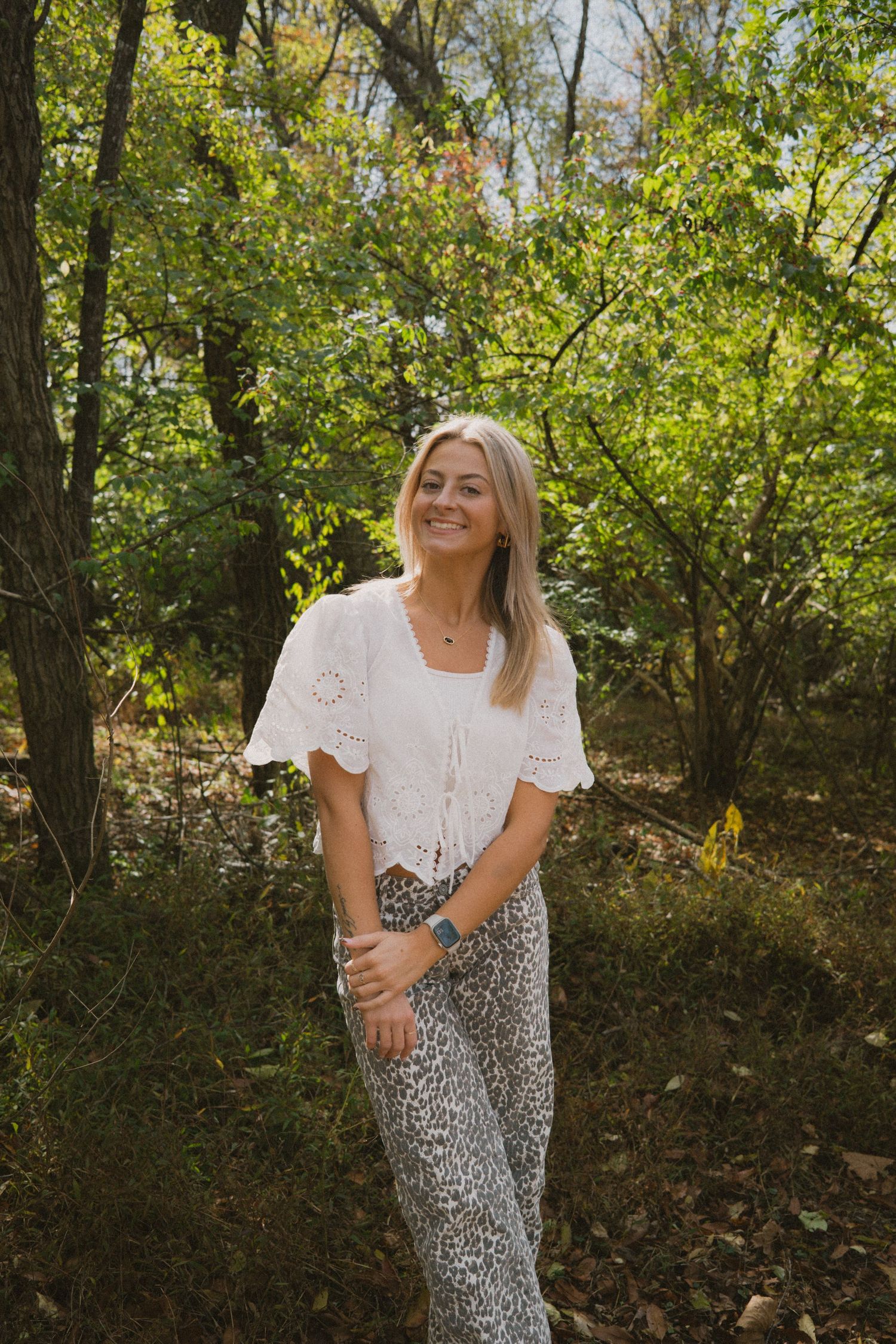 Person in a white eyelet top and leopard print pants poses playfully in a wooded outdoor setting.