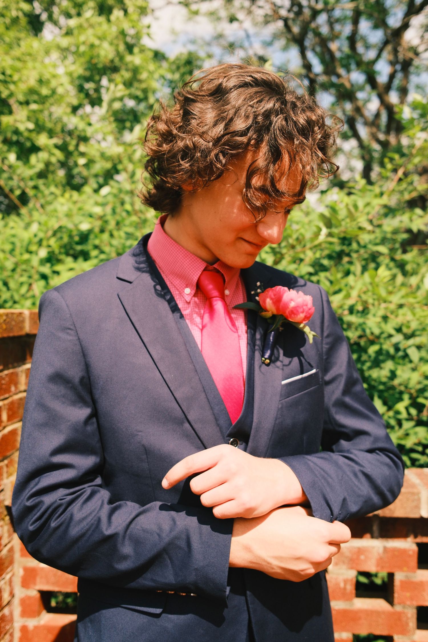 A person in a navy suit and pink shirt adjusts their sleeve cuff while standing outdoors against green foliage.