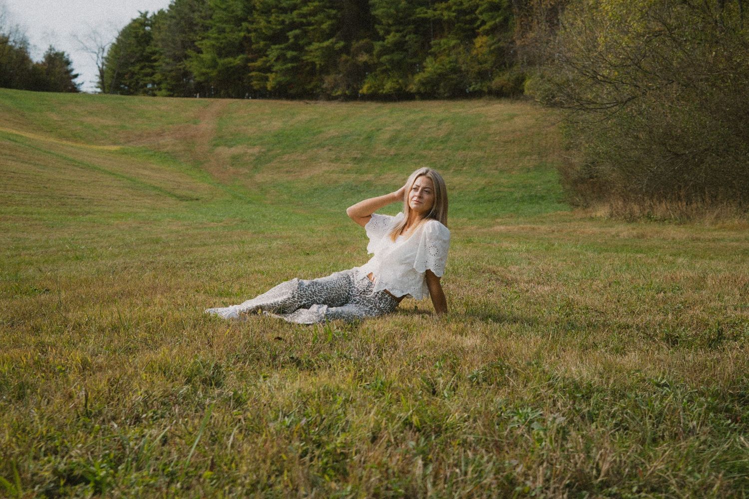 A person in a white dress reclines on a grassy hillside surrounded by evergreen trees on an autumn afternoon.