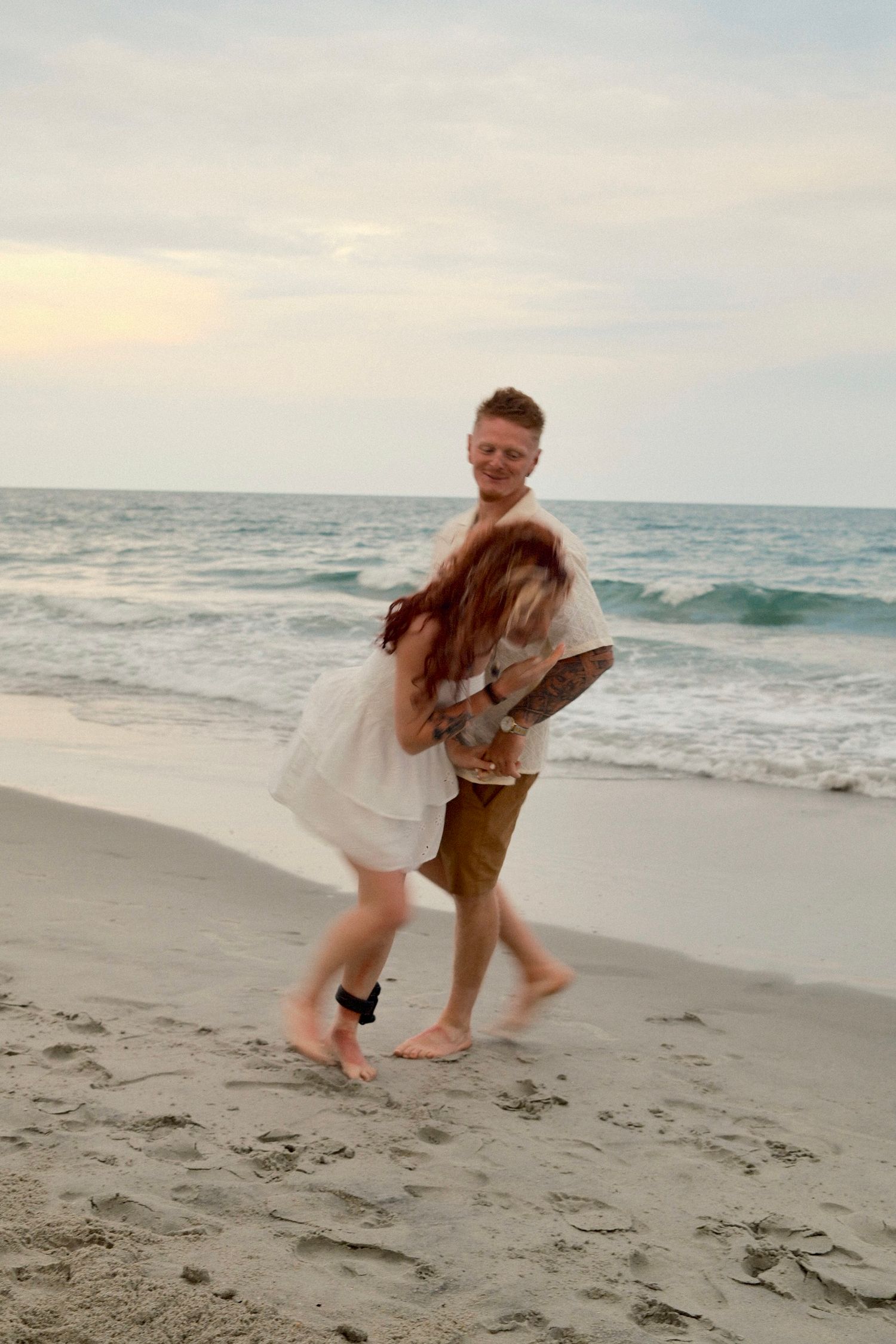 A couple dances playfully on a sandy beach at sunset with ocean waves in the background.