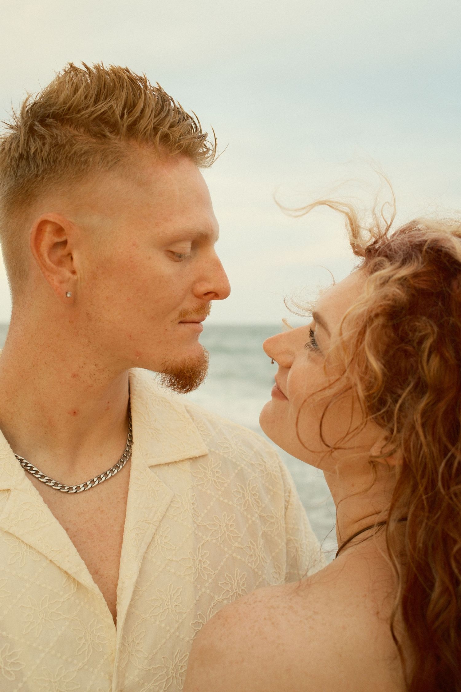 A tender beach photoshoot showing a couple in white clothing against a soft ocean backdrop.