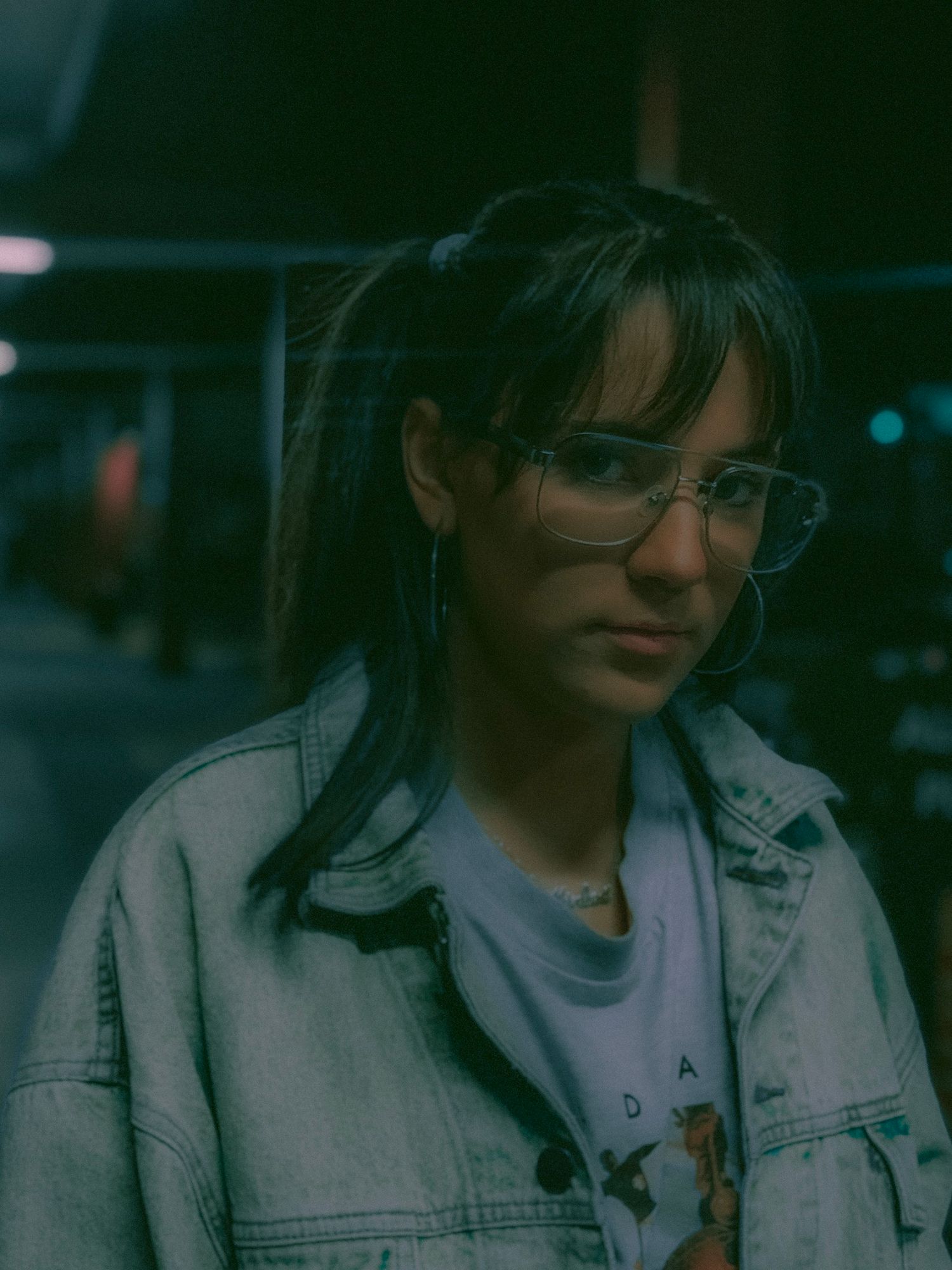 Person wearing glasses and a light colored jacket poses in a moody dark setting with atmospheric lighting.