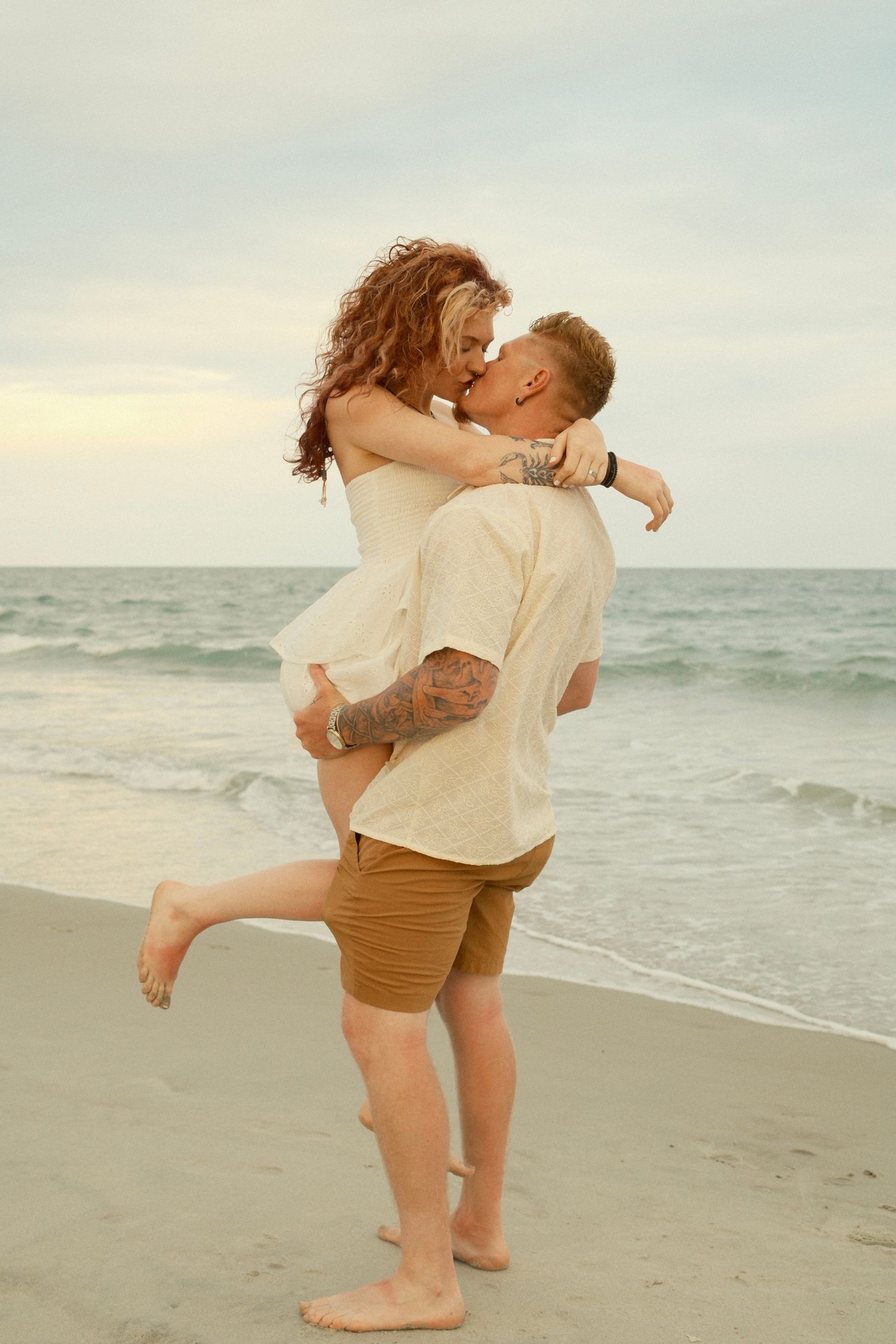A playful couple shares joyful moments on a sandy beach with ocean waves in the background.