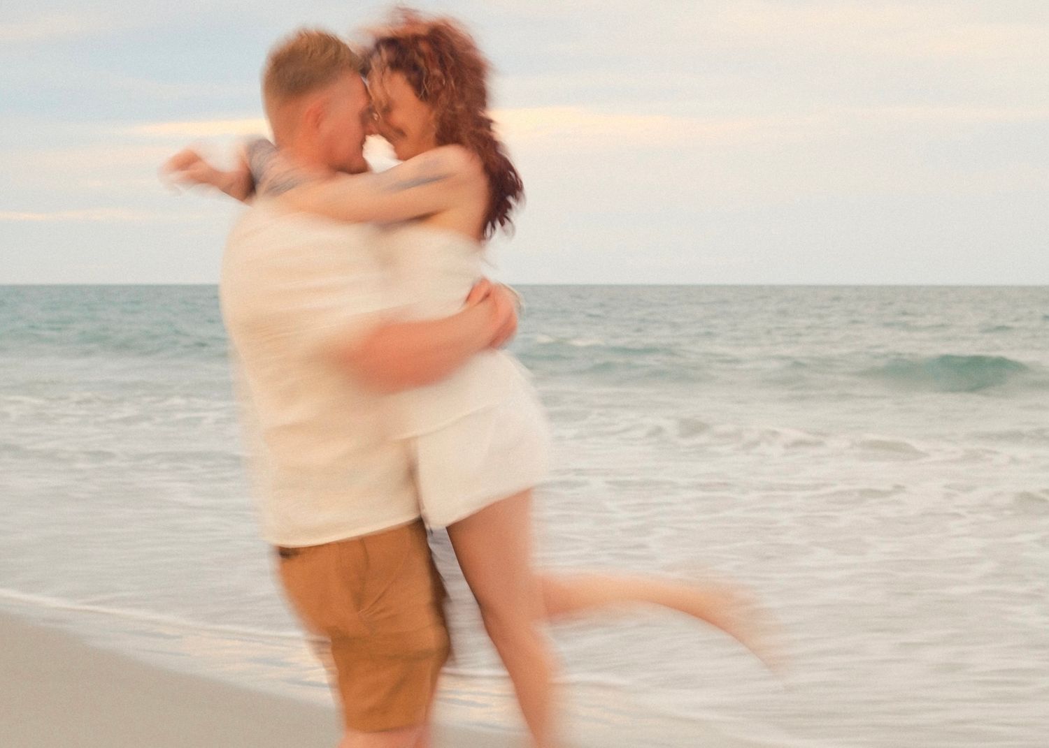A couple embraces on a sunny beach with ocean waves in the background.