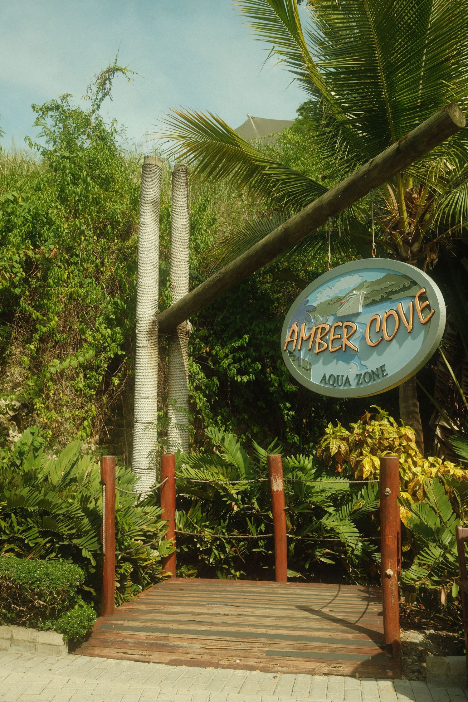 Wooden walkway entrance with Amber Cove and Aqua Zone signs surrounded by tropical palm trees.